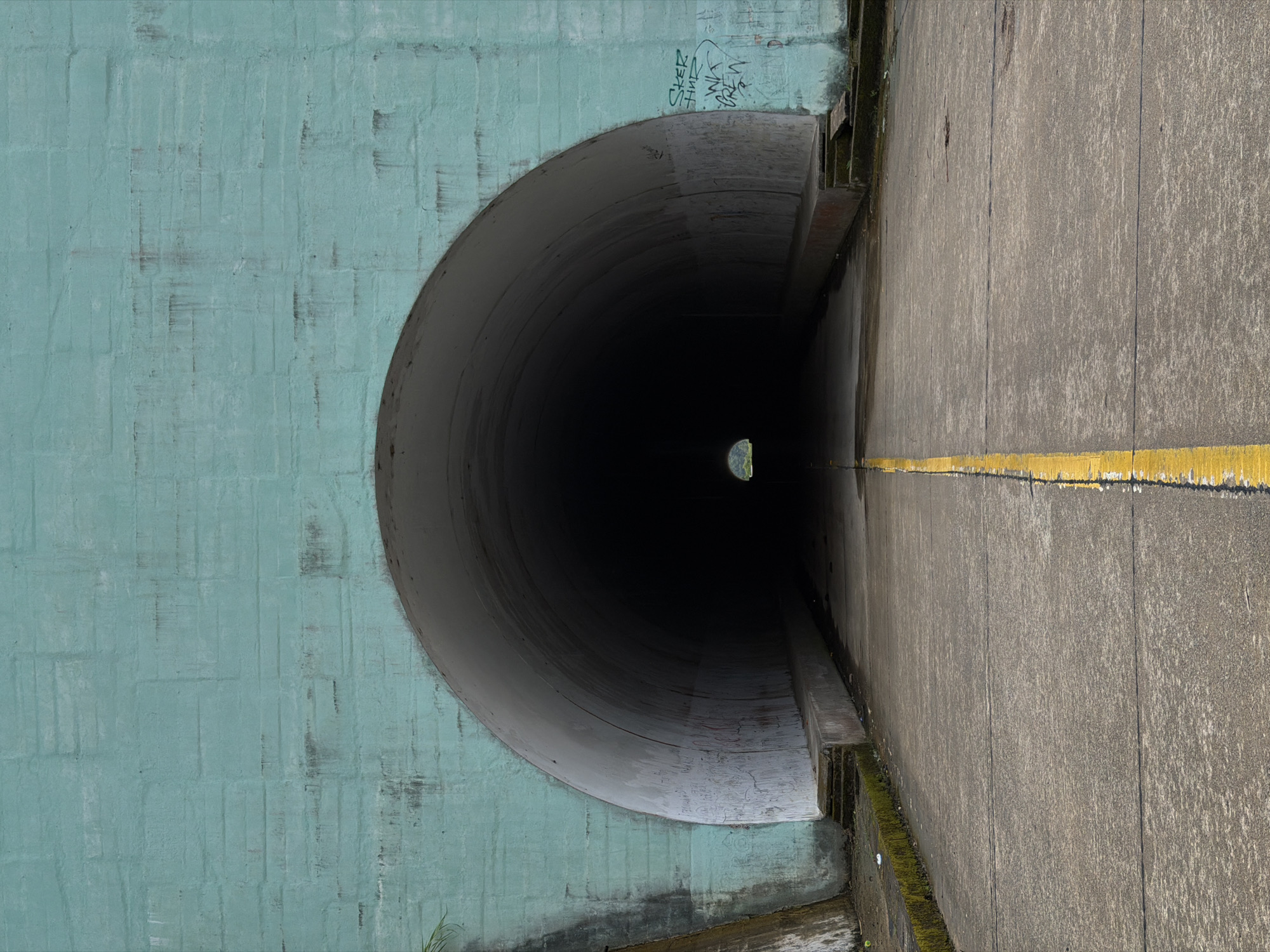The image depicts a section of a tunnel or culvert located in Huế, Vietnam. The entrance to the tunnel is a large, circular opening in a teal-painted brick wall. The interior of the tunnel is dark, with a small opening visible at the far end, suggesting daylight. To the right of the tunnel entrance is a concrete walkway or road with a yellow line marking. The wall adjacent to the tunnel opening features graffiti. Moss and vegetation are present at the base of the concrete walkway. The scene suggests an urban or industrial area.