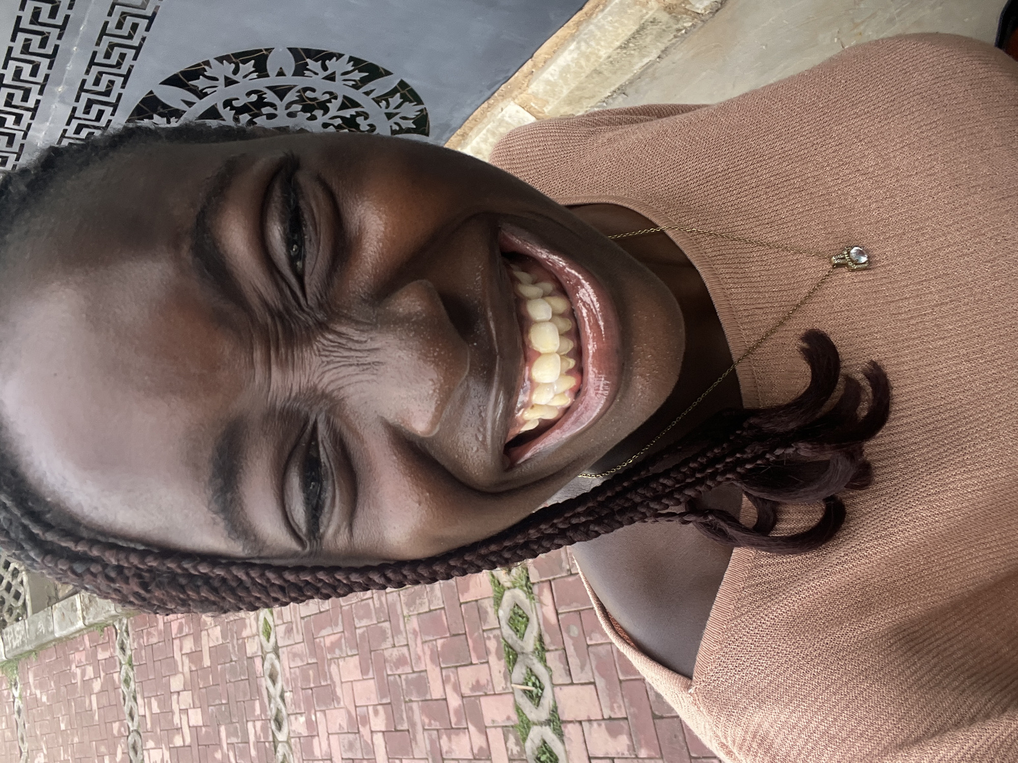 A close-up, frontal view of a young Black woman's face, captured from the shoulders up, reveals her broad, toothy smile. Her eyes are crinkled in mirth, and her expression is lively. She has dark, braided hair that falls over her left shoulder, and she is wearing a light peach-colored knitted top with a delicate gold necklace featuring a small pendant. The background shows a textured, grey wall with a geometric design and a section of red brick paving, suggesting an outdoor urban setting. The lighting indicates it is daytime. While the image doesn't explicitly state the location as Karum, Nigeria, the context implies an urban environment within the country.