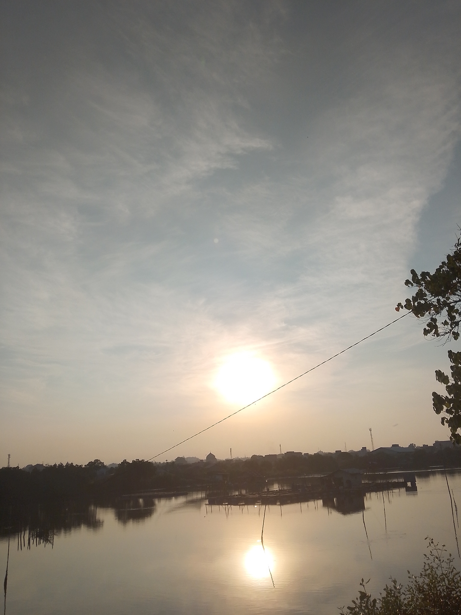 The image depicts a serene waterfront landscape at either sunrise or sunset, characterized by a warm, golden glow. The sun, a brilliant disc, is positioned in the lower-left quadrant of the sky, casting a strong reflection that stretches across the still water surface towards the viewer. The sky is a blend of pale blue and grey, adorned with wispy, streaky clouds that suggest fair weather with some atmospheric haze.
A dark utility wire runs diagonally across the upper and middle portions of the image, starting from the upper left and extending towards the mid-right. To the far right, a silhouette of tree branches with visible foliage partially frames the scene.
In the distance, a low-lying horizon shows a strip of land with dark silhouettes of numerous buildings and dense vegetation, suggesting a town or village along the water's edge in Lhokseumawe, Indonesia. Among these distant structures, details in the cropped views hint at the dome of a mosque and several communication towers. On the water itself, nearer to the distant bank, several dark, rectangular floating structures are visible, possibly indicating fish farms or small riverside dwellings, all contributing to the reflections on the calm surface. On the closer left bank, several vertical poles or stakes rise from the water.
The overall scene conveys a sense of peacefulness and natural beauty, capturing a quiet moment in a coastal or riverine environment during the golden hour. No visible text is present in the image.