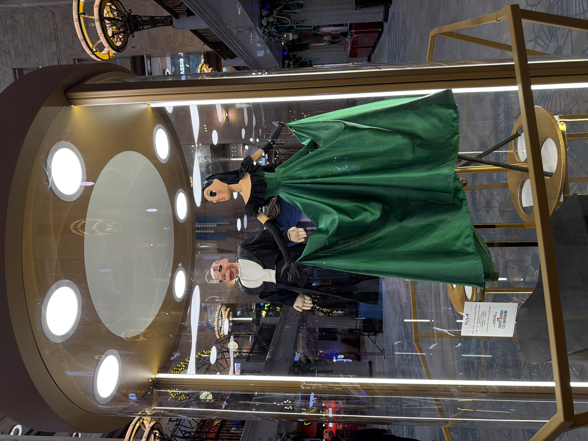 The image depicts a puppet display in a glass case. The case includes two puppets, one of a woman in a green dress and another of a man in a black suit. The display case features a clock-like structure with illuminated round sections. The surrounding environment appears to be an indoor public space, possibly a mall or a museum, based on the architectural elements and visible shops. Based on the text on the sign inside the case, it is likely that the image was taken in Moscow, Russia.