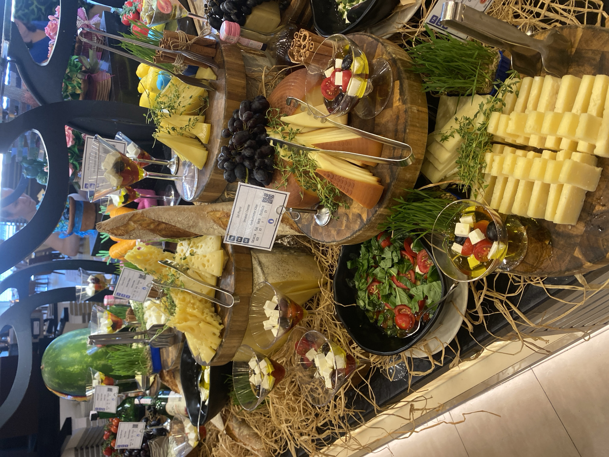 This image depicts a buffet display, likely in a hotel or restaurant, featuring a variety of cheeses, bread, fruits, and vegetables. The overall presentation suggests a breakfast or brunch spread. The specific mention of Aksu, Turkey, places the scene in this city, which is known for its agricultural production and tourist attractions.
Detailed description of the scene:
The foreground of the image is dominated by a rustic buffet setup. Several wooden platters are stacked and arranged, showcasing an assortment of cheeses. Some cheeses are sliced into neat portions, while others are presented as larger wedges or wheels. The cheese varieties appear to include hard, semi-hard, and possibly some softer types, with colors ranging from pale yellow to a deeper orange.
Scattered amongst the cheeses are sprigs of fresh herbs, likely for garnish and aroma. Alongside the cheese, there are bowls of olives, tomatoes, and what appears to be feta cheese marinated in olive oil. A large loaf of crusty bread, cut lengthwise, is prominently displayed on one of the platters.
In the background, a large green watermelon is visible, suggesting the availability of fresh fruit. Several martini glasses are filled with a mixture of cubed cheese, cherry tomatoes, and olives, presented as individual appetizers. Bunches of dark grapes are also part of the display, adding a touch of color and richness.
The buffet is arranged on a dark countertop, with decorative straw elements providing a natural, textured base. Behind the buffet, glimpses of other buffet items, such as bottled beverages and possibly more produce, can be seen. In the far background, out of focus, are patrons of the establishment, some of whom appear to be selecting food. The lighting suggests an indoor setting, possibly with natural light filtering in from windows. The overall impression is one of abundance and a well-curated food selection.
Considering the context of Aksu, Turkey, this buffet likely reflects the local culinary offerings, potentially incorporating regional cheeses and fresh produce common to the area. Aksu is situated in the Antalya province, a region known for its Mediterranean climate, which supports diverse agricultural activities.