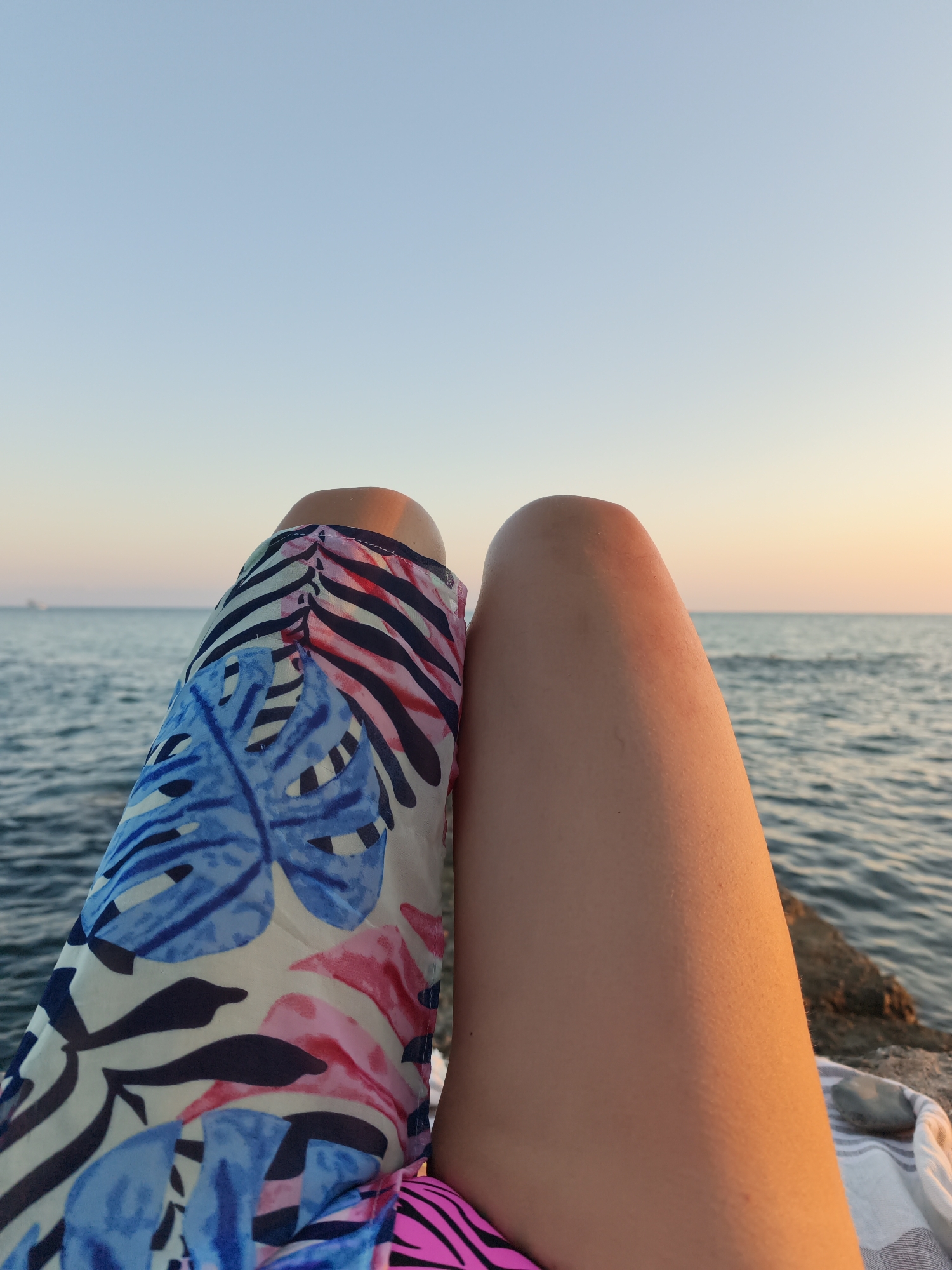 The image shows a close-up view of a person's legs against a backdrop of the sea and sky. The person is wearing a colorful sarong with a tropical leaf print in shades of blue, pink, and black, as well as a striped bikini bottom. The legs are positioned to the right of the sarong, with the skin illuminated by a warm, golden light, suggesting it is either sunrise or sunset. The sea is visible in the background, with gentle ripples on its surface. The sky above is a gradient of pale blue, transitioning to a soft orange and yellow near the horizon, indicating the time of day. The scene appears to be outdoors, possibly on a rocky embankment or pier, with a striped towel and a stone visible in the lower right corner. The photo is taken from a low angle, focusing on the lower body of the person and the serene natural environment. The location is not specified in the image, nor is there any indication that it is in Sochi, Russia.
