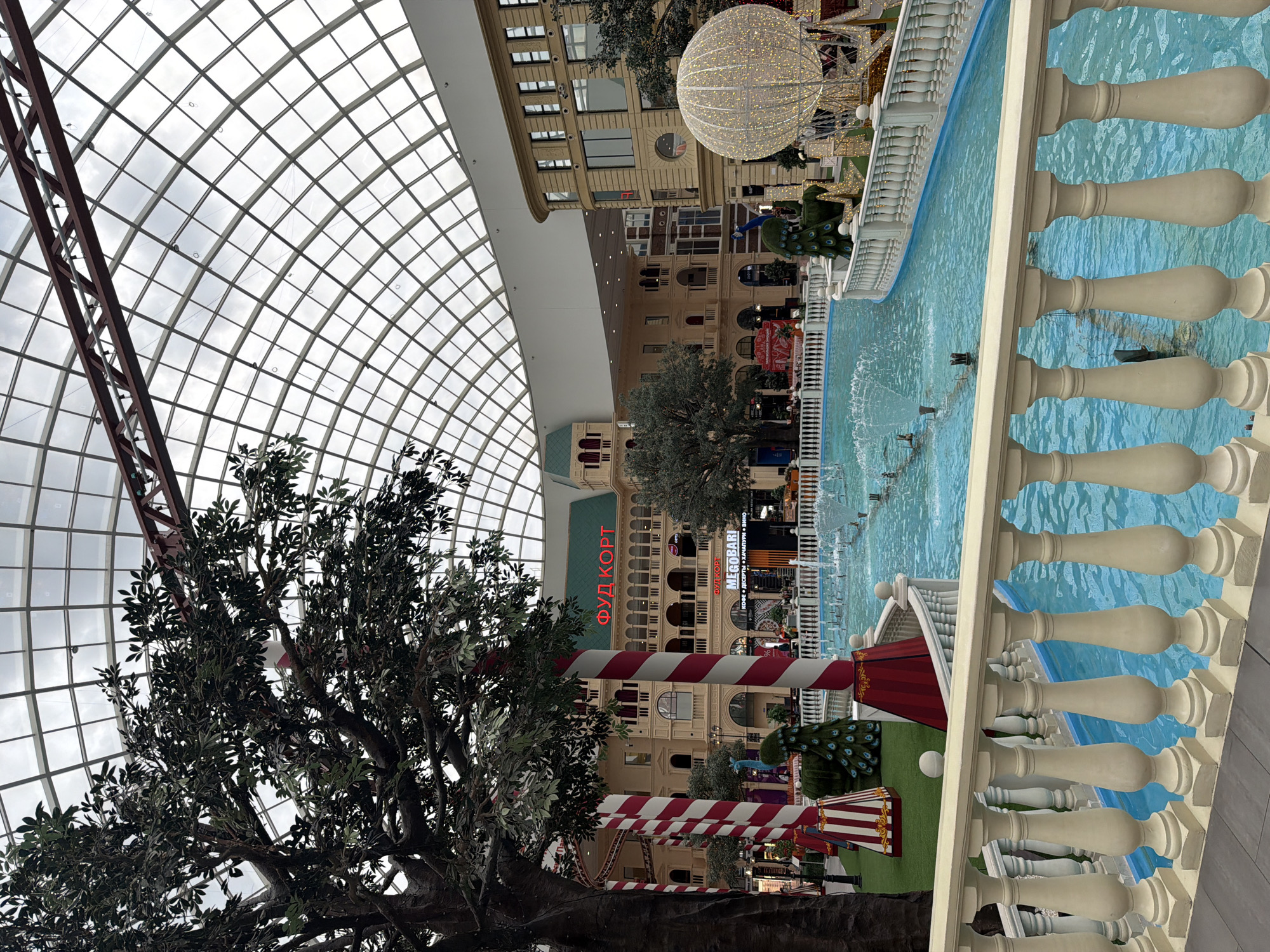 This is a wide-angle, eye-level shot of an indoor atrium resembling a European town square, bathed in natural light from a large, curved glass ceiling. In the foreground, a white balustrade with ornate balusters overlooks a bright turquoise pool with a fountain spraying water. To the left, a large, artificial tree with dark green leaves stands prominently. Several red and white striped columns, reminiscent of circus tents, are positioned near the tree and the pool.
In the midground, a body of water functions as a canal or river, with a fountain creating a visible spray. Beyond the water, buildings with a European architectural style are visible, featuring cream-colored facades, arched windows, and red tiled roofs. One building has large red lettering that reads "ФУД КОРТ" (Food Court) above a green awning. Another section of the building displays "MEGOBAРI" in white text on a black background, with additional Russian text below. Decoratively lit Christmas trees and a peacock sculpture are placed along the canal. To the far right, a large, illuminated sphere, resembling a hot air balloon or Christmas ornament, is visible.
The background consists of the arched glass ceiling structure and more buildings within the vast indoor space. A few scattered individuals are visible in the distance, primarily in the midground near the buildings and along the edge of the water. The overall atmosphere suggests a festive or recreational indoor environment, possibly a shopping mall or entertainment complex.