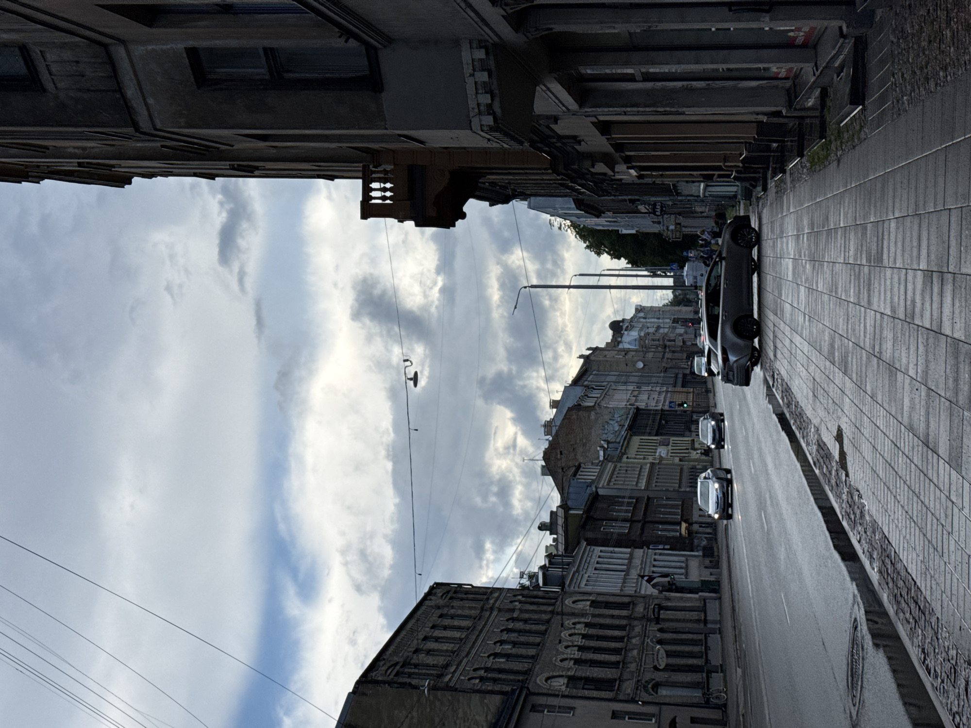 This is a street-level, eye-level, medium shot of a cloudy day in Riga, Latvia. The scene is dominated by a wide, wet asphalt road with a sidewalk paved with grey bricks. Several cars are driving on the road, including a light-colored SUV in the foreground, and a silver sedan with its headlights on. On the left side of the street, there are several old, multi-story buildings with ornate architectural details. The buildings are made of brick and stone, with arched windows and balconies. On the right side of the street, there are more buildings with a similar architectural style. The sky is filled with dramatic, dark clouds, suggesting an overcast or stormy day. There are also visible power lines crisscrossing the sky. The overall atmosphere is one of urban life under a moody sky.