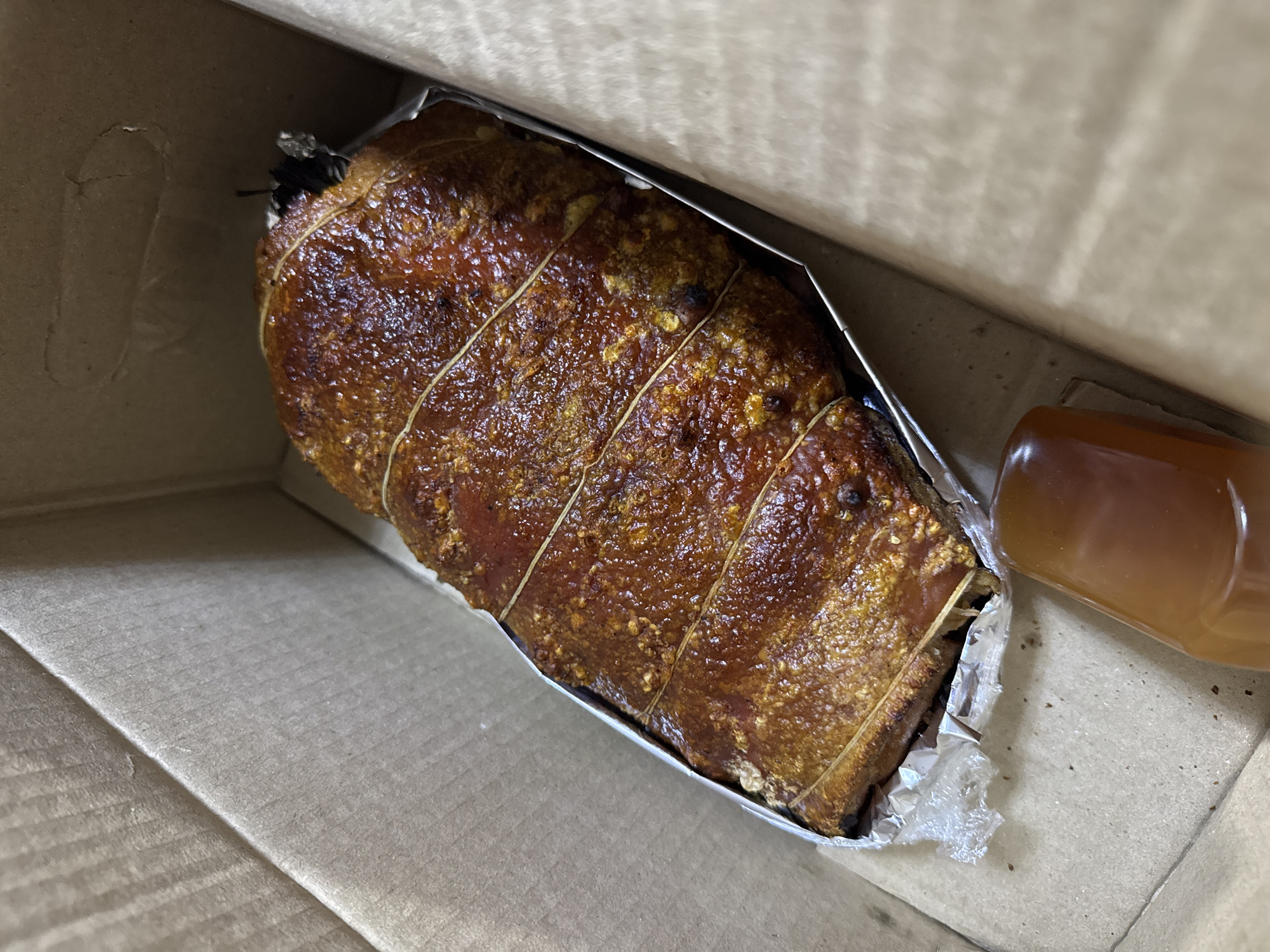 A close-up, high-angle shot depicts a lechon roll secured with string in a corrugated cardboard box lined with aluminum foil. Beside the lechon roll is a small, clear, plastic container filled with a light-brown liquid, possibly gravy or sauce. The lechon roll appears to be resting on a bed of its drippings, which are contained by the aluminum foil. The photograph was taken in Davao City, Philippines.