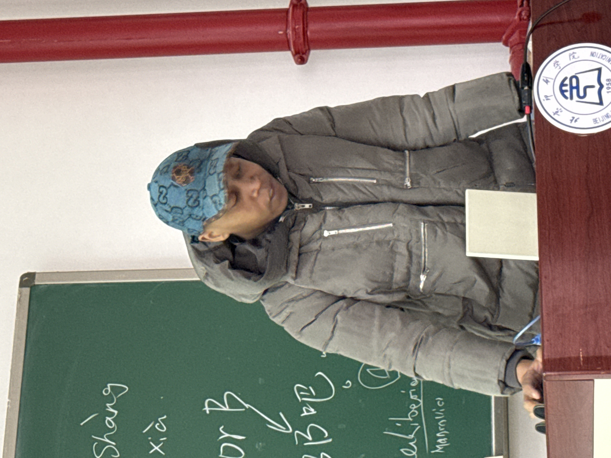 A person with a blue patterned cap is at a podium near a chalkboard. They are wearing a gray puffer jacket. The chalkboard has Chinese characters and English writing on it, including "Dr B," "Manrollici," and "Liberia." A red pipe is visible near the ceiling. A sticker on the side of the podium reads "BEIJING." The setting is presumably in the city of Daxing District, China.