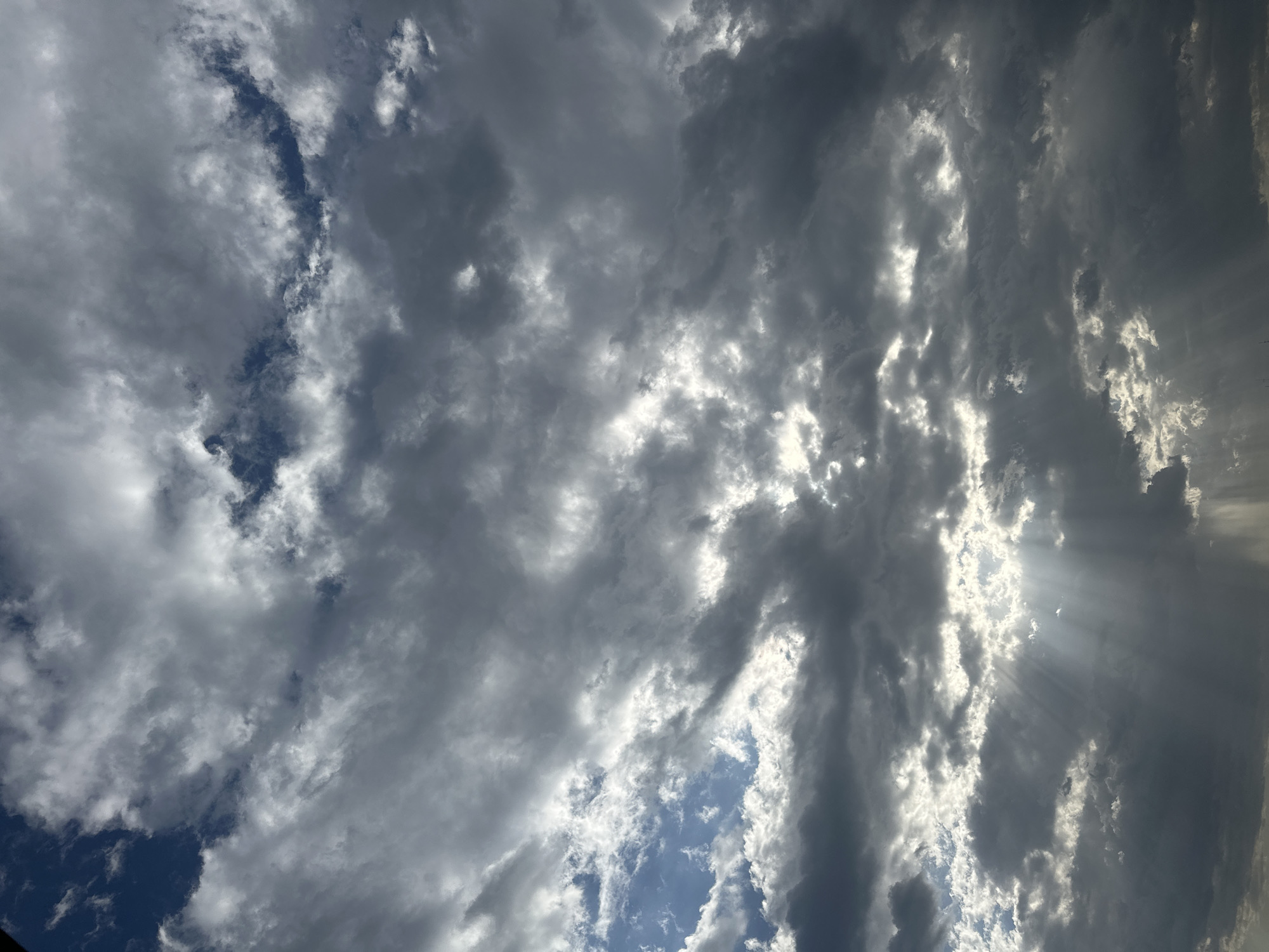 The image is a low-angle shot of a cloudy sky. The clouds are mostly gray and white, with some patches of blue sky visible in between. In the lower left, a small section of a dark object is visible in the lower-left corner. Sunlight is filtering through the clouds, creating a dramatic effect with rays of light visible on the right side of the frame. There are no people or objects present in the scene. The location is unknown.