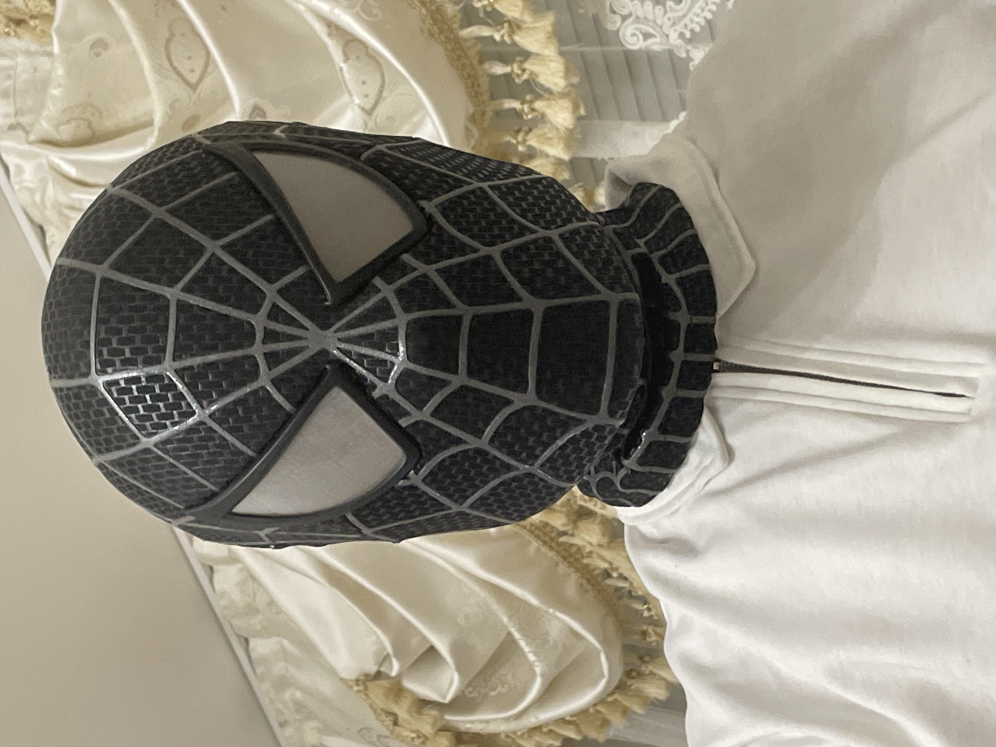 The image primarily features a black and gray Spider-Man mask, resembling the Symbiote suit, positioned upright. The mask has a textured, grid-like pattern in black, overlaid with prominent silver web-like lines. It features large, distinct white eye lenses.
The mask is resting on or in close proximity to a plain white garment, possibly a t-shirt or hoodie, with a visible zipper pull near what appears to be the neck or collar area. In the background, there is a light-colored, possibly cream or beige, fabric with subtle patterns and a satiny finish, adorned with decorative tassels or fringe. This background fabric suggests a soft furnishing like a curtain or tablecloth.
The scene is static, with no activity or event taking place. The lighting is soft and even, indicating an indoor setting during what appears to be daytime. No people are visible, and no emotions are conveyed. There is no visible text in the image. While the location context provided is Ihtiman, Bulgaria, there are no specific visual cues within the image itself to confirm this geographical location.