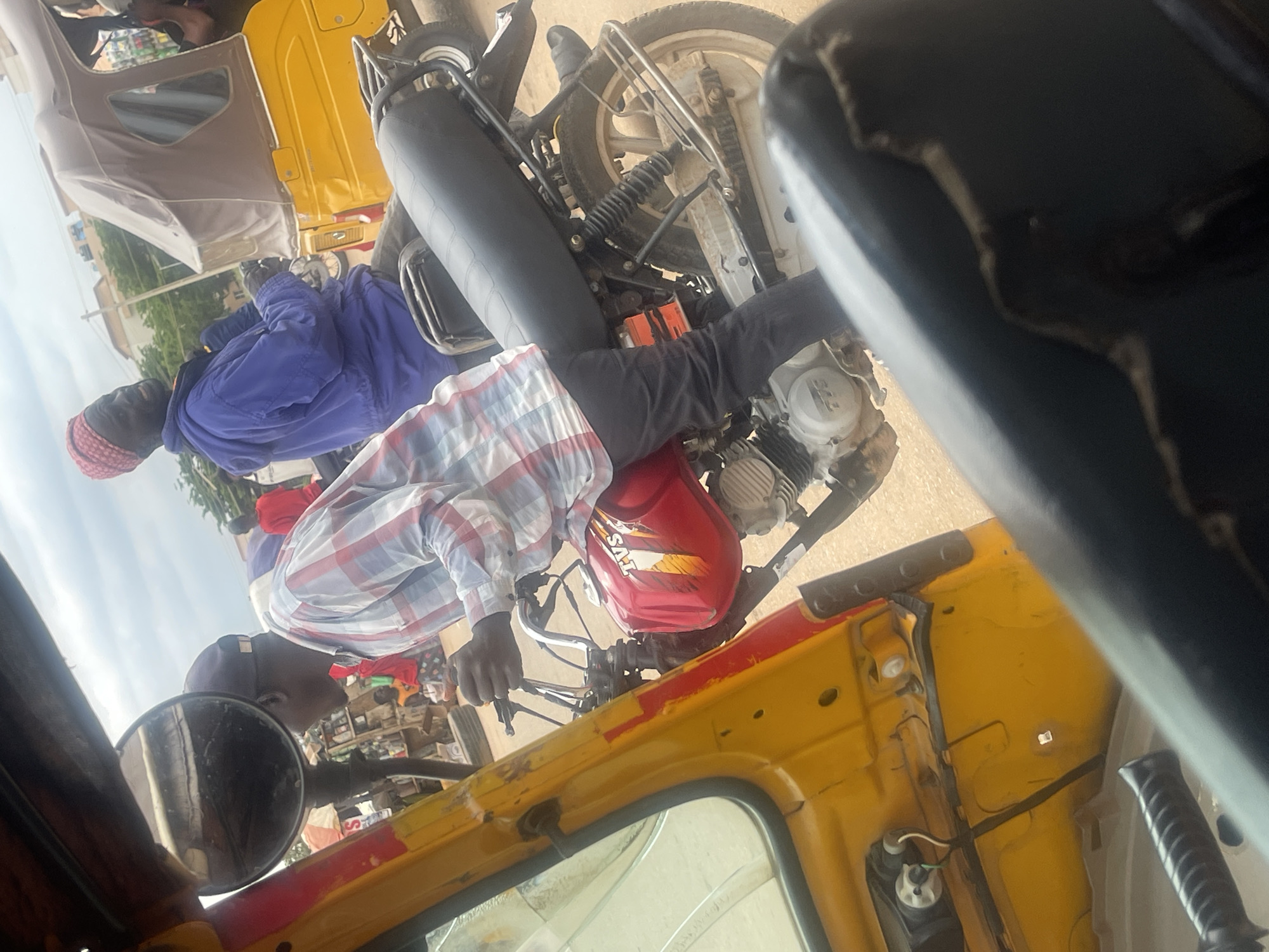 The image depicts a street scene in Galadimawa Village, Nigeria. Three people are visible, two men and a woman. One man is driving a yellow motorized tricycle, also known as an "Okada" or "Keke Napep." He is wearing a red and white checkered shirt and a dark head covering. Seated behind him is a woman wearing a purple jacket and a red headscarf. Another man, possibly a passenger, is visible in the foreground, wearing a dark head covering and a red and white checkered shirt. The tricycle has a yellow body with some red detailing and a black seat. The background shows a blur of activity, with buildings and trees suggesting an urban or semi-urban environment. The lighting indicates it's daytime. The focus is on the people and the vehicle, suggesting a moment of transportation or travel. There is no discernible text in the image.