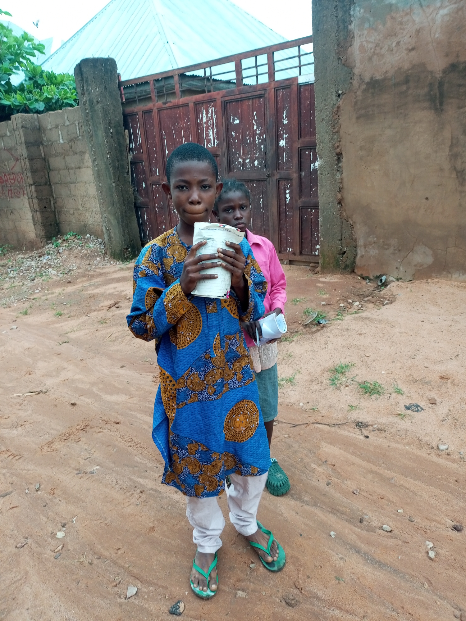 In Tempure, Nigeria, a young boy stands in front of a weathered, reddish-brown metal gate. He wears a blue and gold patterned traditional garment and white pants, accessorized with green flip-flops. He holds a rolled-up notebook in his hands. Behind him, a young girl, partially obscured, wears a pink shirt and blue shorts. The ground is a sandy, reddish-brown dirt path with scattered stones and small patches of dry grass. A rough, textured concrete wall forms the right side of the frame, with a large, weathered concrete pillar to the left, adjacent to the gate. A metal fence with horizontal bars is visible above the gate, and a light-colored, corrugated metal roof structure is partially visible at the top of the image. The overall environment suggests a rural or semi-urban setting with modest construction.