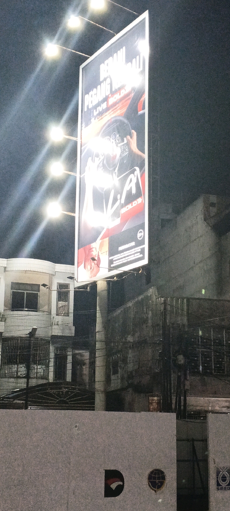 An outdoor billboard at night is illuminated by overhead lights in Medan, Indonesia. The billboard displays an advertisement with large text that reads "BERANI PEGANG KENDALI" and "LIVE BOLD". A person's hands are shown gripping a steering wheel, and a logo with "A" and "BOLD" is also visible. The billboard is positioned in front of a multi-story building with windows and balconies. Another building with a textured, darker facade is to the right of the billboard. In the foreground, a low, light-colored wall displays several logos, including one resembling an eagle's head and another depicting a globe. The overall scene suggests an urban setting at night.