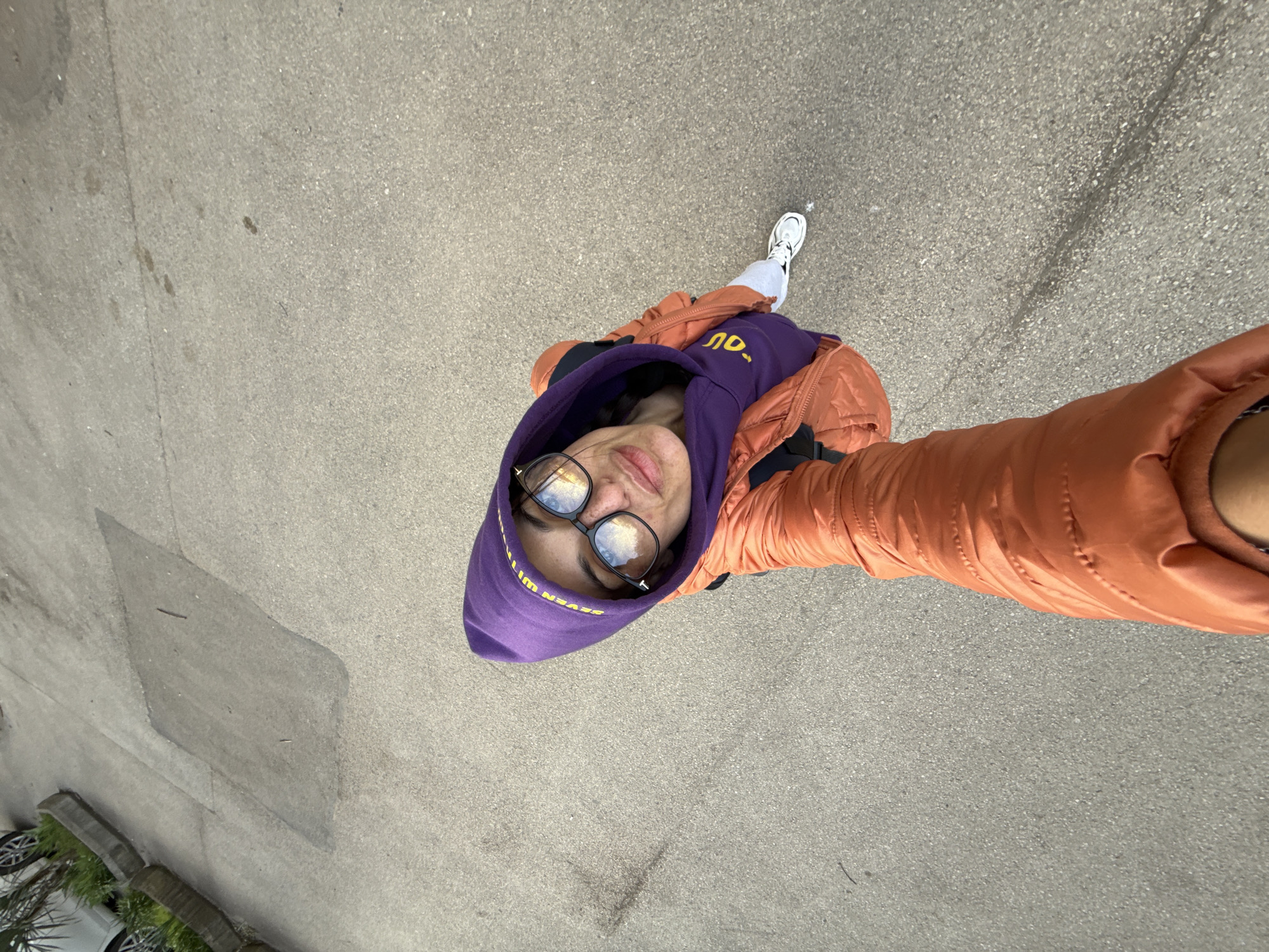 A high-angle shot depicts a person with fair skin wearing glasses, a purple hooded sweatshirt, and an orange puffer jacket. The person is holding the camera at arm's length, creating a selfie. The sweatshirt has yellow lettering visible. The person is standing on a gray asphalt surface, likely a street or sidewalk.
In the lower left corner of the frame, there is a partial view of parked cars and greenery.
It is not possible to verify the specific location as the city of Pleven, Bulgaria based on the visual information in this image.