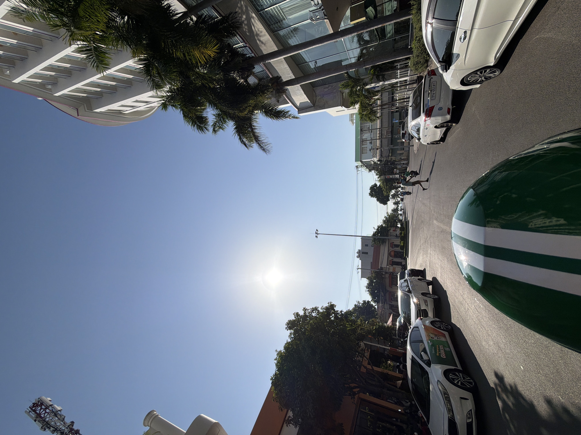 The image depicts a street scene in Nha Trang, Vietnam, on a bright, sunny day. The viewpoint is from the perspective of someone on a motorcycle, looking forward and slightly upwards.
In the foreground, the front of a green and white striped object, likely a motorcycle helmet or a carrier, is visible.
The street is paved and appears to be a two-way road with multiple lanes. Several cars are parked and driving on the road. On the left side of the street, a white sedan with a green and orange sticker is parked. Further down the street, a white minivan and another dark-colored car are visible. On the right side of the street, a white sedan is parked, and another white sedan is driving away.
Buildings line both sides of the street. On the right, a modern, curved white building with many windows and balconies is prominent, with tall palm trees in front of it. On the left, there are lower buildings, one with an orange facade and trees casting shadows on the street.
The sky is clear and blue, with the sun shining brightly, causing some lens flare. There are power lines and streetlights visible. In the distance, people can be seen walking and riding motorcycles. The overall impression is a typical urban street in a tropical climate.