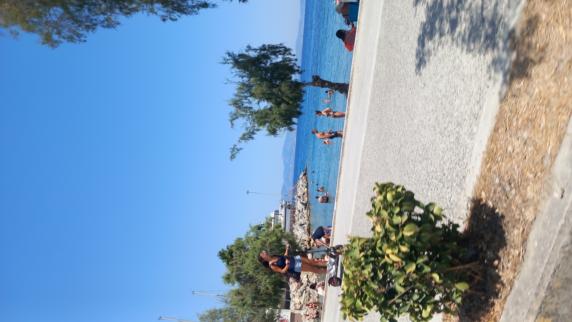 A woman is riding an electric scooter on a paved road next to a beach. She is wearing a dark blue tank top and denim shorts. She has a backpack slung over her left shoulder. In the background, people are swimming and relaxing in the ocean. There are trees and boats visible on the water. The sky is clear and blue. The location is near the city of Heraklion, Crete, Greece, likely at a beach accessible by road. The scene depicts a sunny day at the seaside, with a mix of recreational activities.