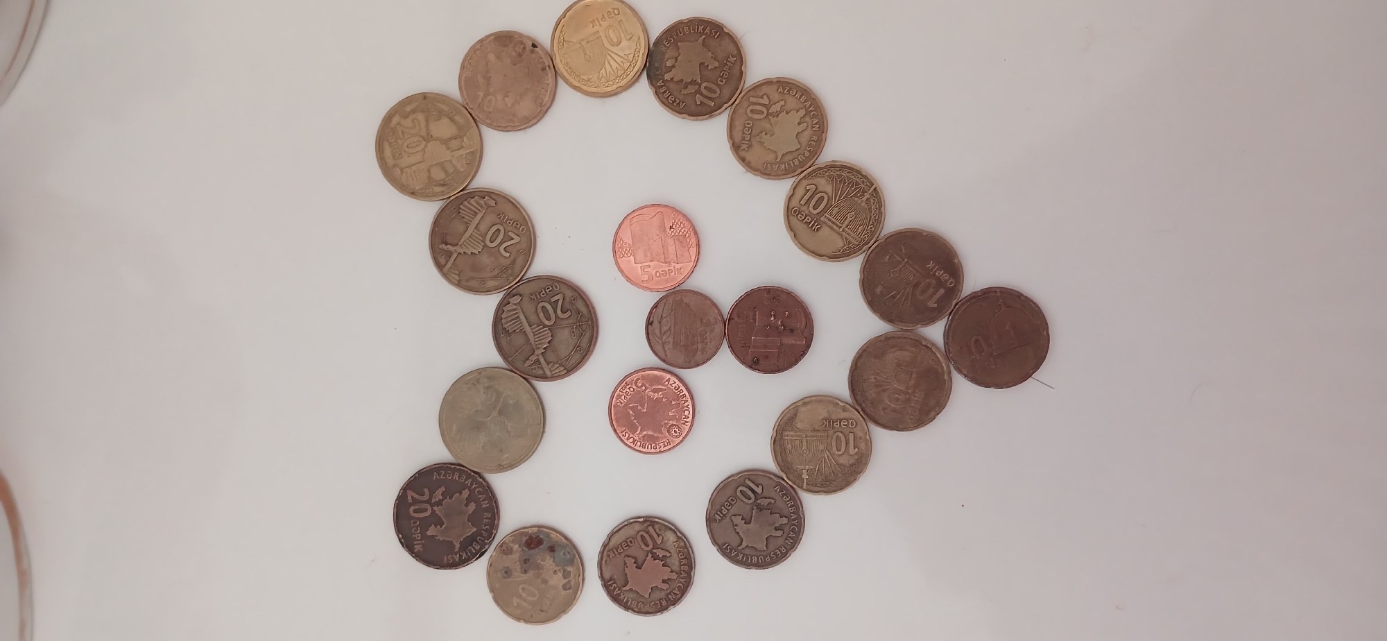 This image displays a collection of Azerbaijani coins arranged on a white surface, forming a loose circular or heart-like shape. The coins are of various denominations, including 5, 10, and 20 qəpik. Some coins are copper-colored, while others appear to be a brass or silver alloy. The visible text on the coins includes "AZƏRBAYCAN RESPUBLİKASI" (Republic of Azerbaijan) and denominations like "10 QƏPİK," "20 QƏPİK," and "5 QƏPİK." Some coins show intricate designs, such as buildings or maps of Azerbaijan. The arrangement of the coins is the primary subject, with no people or animals present. The lighting suggests an indoor setting, likely during the daytime, with no specific weather cues. The overall impression is a still life composition focused on currency.