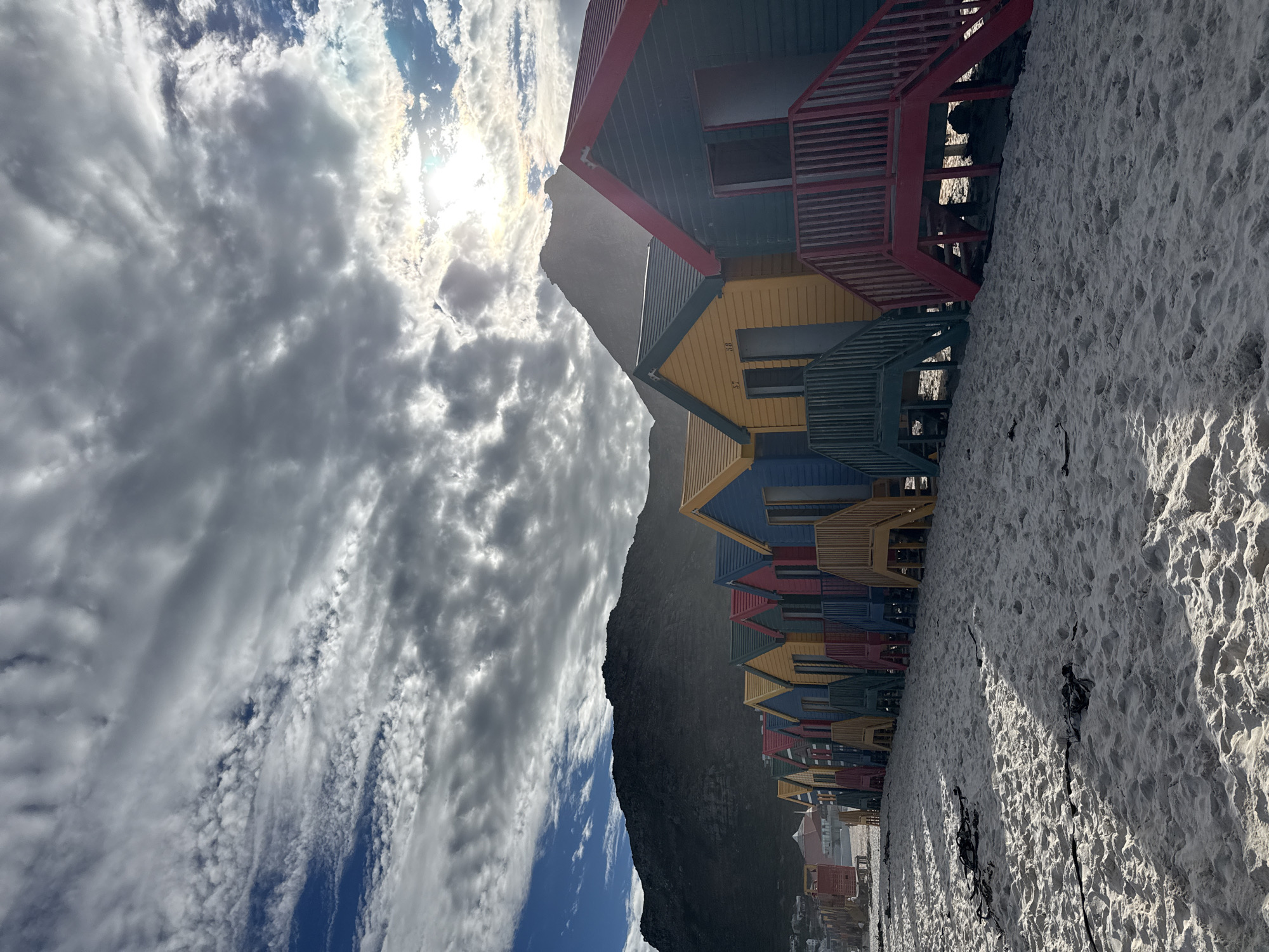 The image shows a row of colorful beach huts at Muizenberg Beach in Cape Town, South Africa. The huts are painted in various colors, including red, yellow, blue, and green. A sandy beach is in the foreground. A dark, rocky mountain is behind the huts, and a cloudy sky with the sun shining through is overhead.