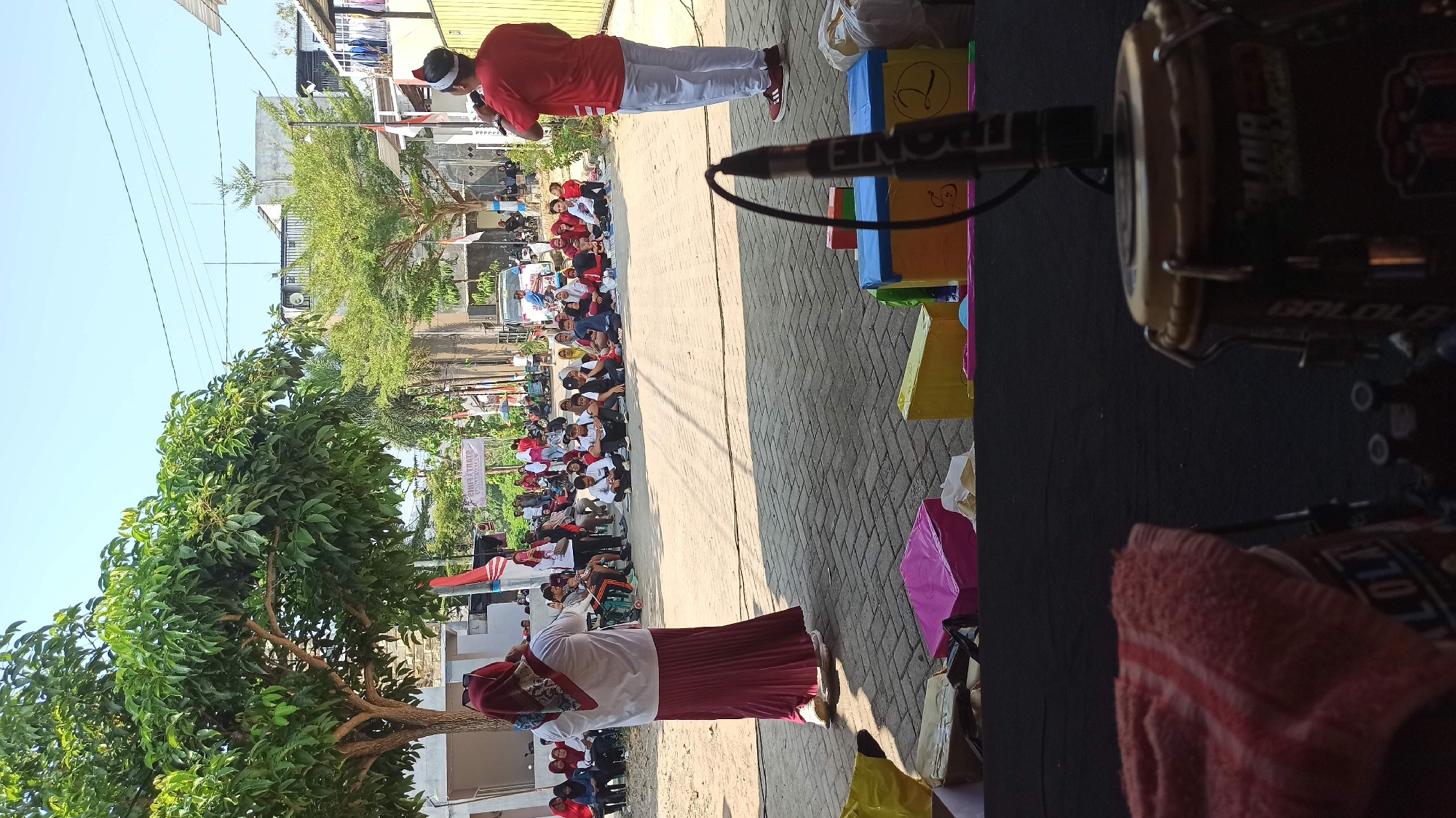 The image depicts an outdoor gathering on a paved surface, likely a street or courtyard, in a residential area. In the foreground, partially visible, is a drum with red fabric draped over it and a microphone stand. The main scene shows a group of people, including children and adults, sitting on the ground facing a woman in a red and white headscarf and skirt, who is speaking or performing. Another man in a red shirt and white pants stands to the right, also facing the seated audience. A banner with Indonesian text is visible in the background, along with trees, buildings, and parked vehicles, suggesting a community event. The atmosphere appears to be casual and celebratory, possibly a local festival or educational activity. The presence of colorful boxes in the middle ground could indicate gifts or props for the event. The lighting suggests it is daytime.