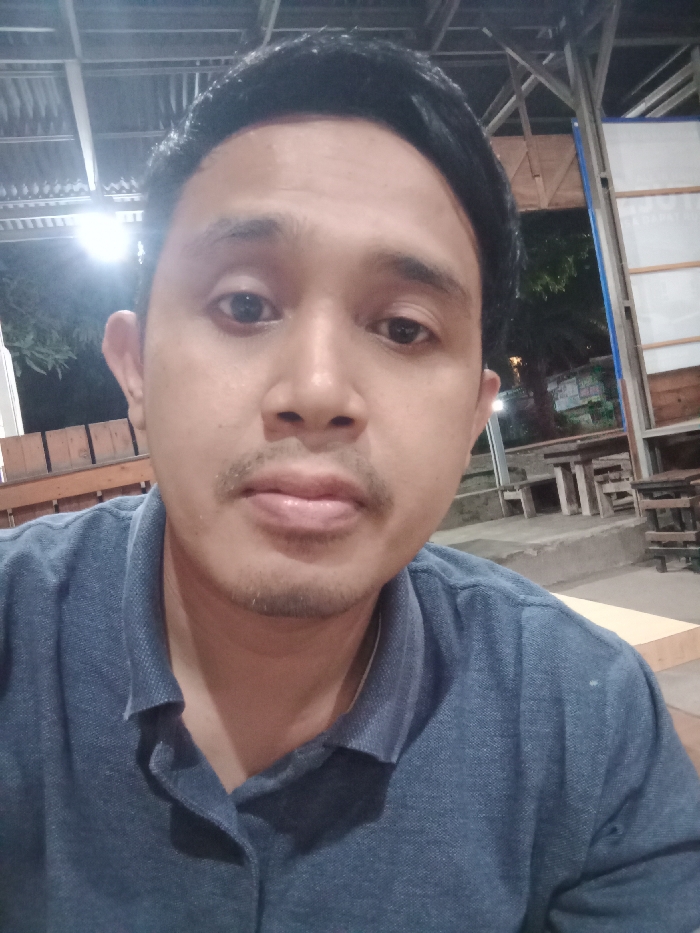 A male individual is depicted in a frontal portrait, centered within an outdoor or semi-open-air commercial establishment at night in Kebonsari, Indonesia. The subject, an adult male with short dark hair, dark eyes, and a light mustache and goatee, wears a dark blue polo shirt and maintains a neutral expression while looking directly into the camera. The background features a corrugated metal roof supported by metal framing, a wooden railing on the left, and a structure with vertical metal beams and white panels on the right. Several rustic wooden tables and benches are visible in the mid-ground, positioned on a concrete platform. Artificial light sources illuminate the scene; a bright overhead light is present on the left, and distant ambient lighting with visible trees and urban structures is evident against the dark night sky.