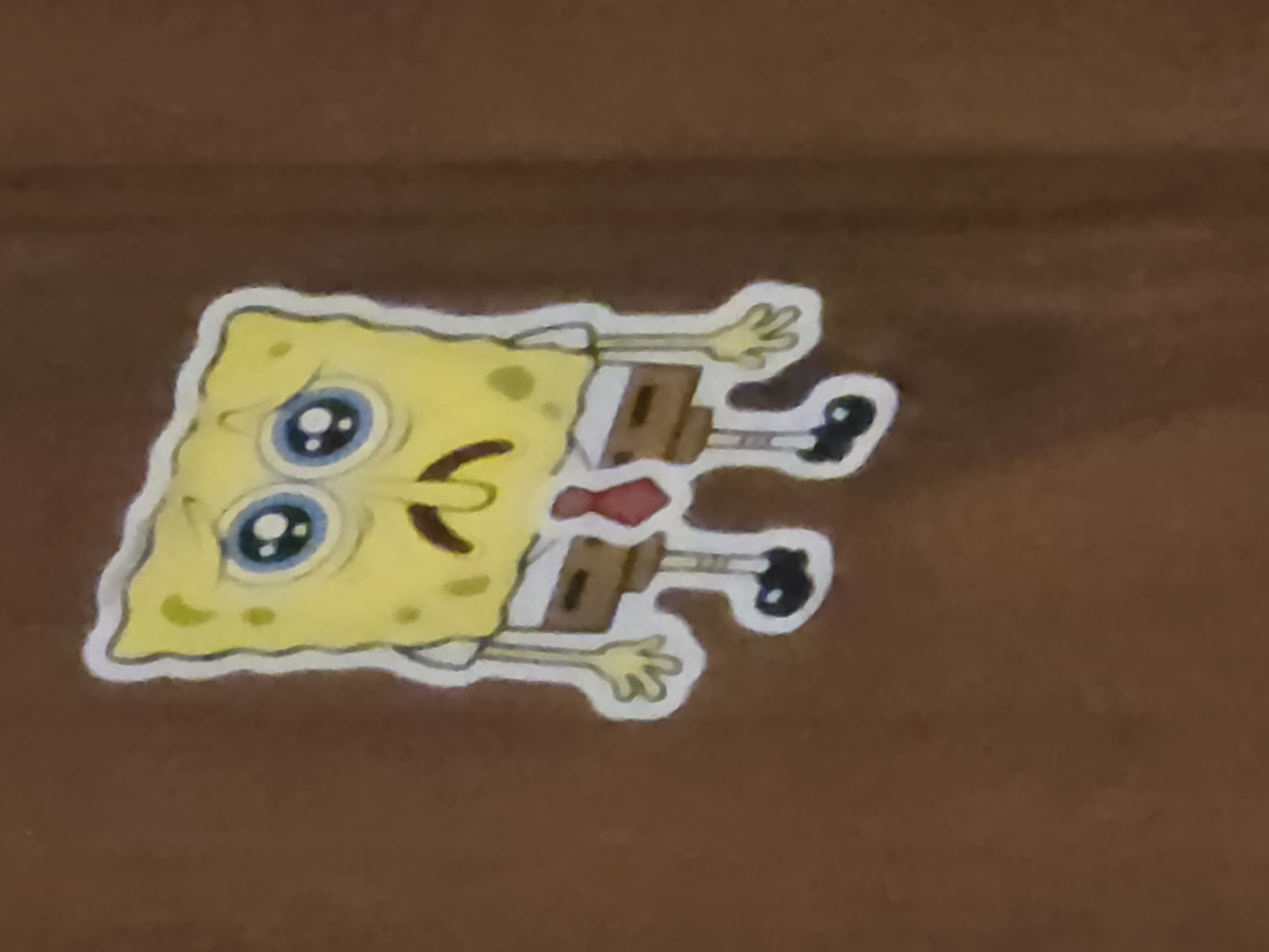 A sticker of SpongeBob SquarePants, depicted with a sad expression, is affixed to a brown surface, possibly a wall or a piece of furniture. The sticker is approximately centered in the frame and its edges are clearly defined by a white border. SpongeBob is in a stylized, cartoonish manner, with his characteristic yellow square body, large blue eyes, and a downturned mouth. He is wearing his typical white shirt, brown square pants, and a red tie. His limbs are extended, and his hands are open, adding to his forlorn appearance. The background is a uniform, muted brown, which contrasts with the bright yellow of the character. The lighting appears to be ambient, casting a soft glow on the sticker and the surrounding surface. The sticker appears to be made of a vinyl or paper material, and its surface is smooth. The overall impression is that of a decorative item placed in an indoor setting.