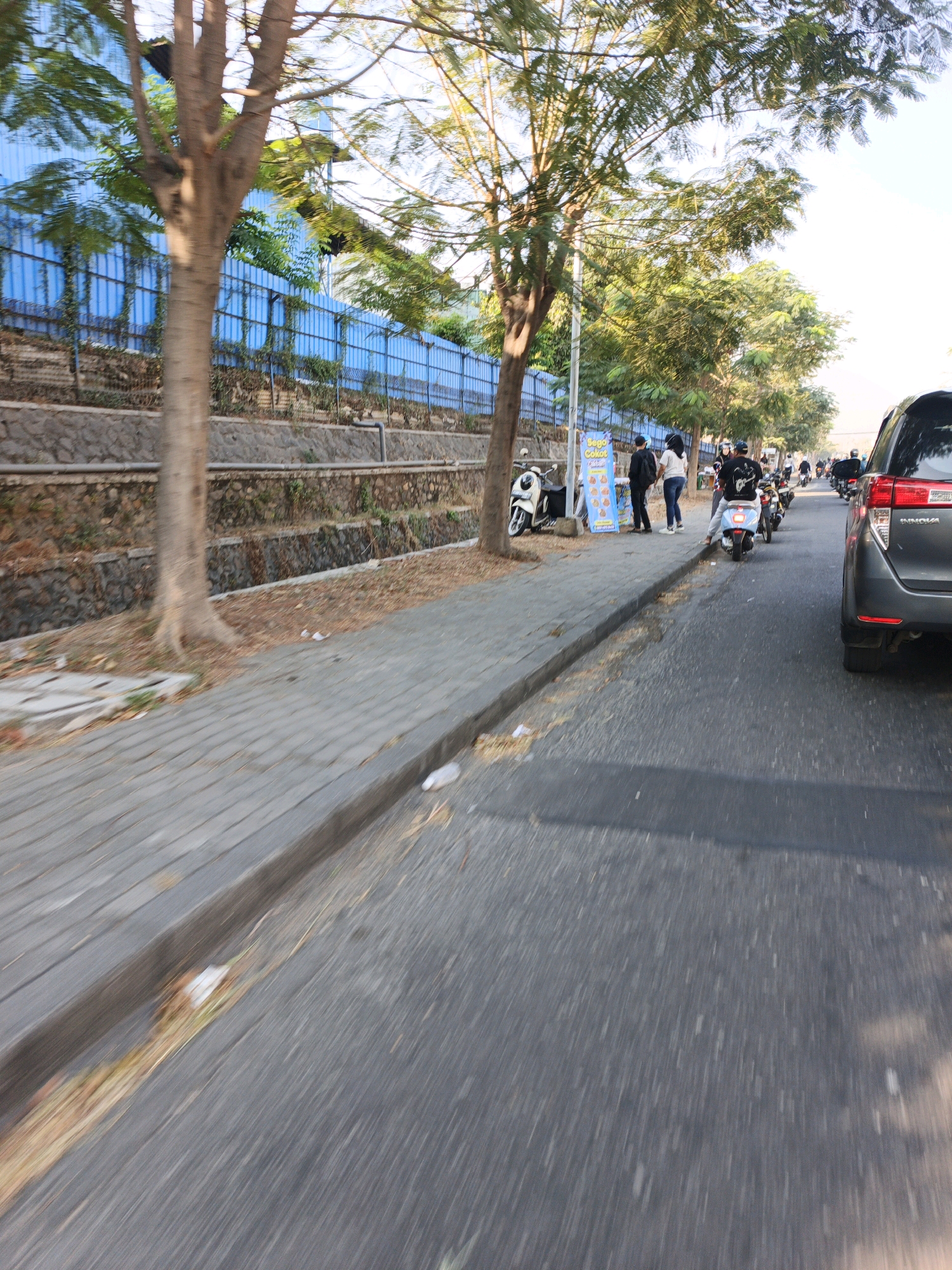 The image captures a street scene in Ngoro, Indonesia, with a focus on a row of parked motorcycles and a car. On the left, a sidewalk is lined with trees and a blue fence. Several motorcycles are parked along the sidewalk, some with riders. A group of people, including two individuals in the foreground standing by a food stall, are visible. A dark-colored car is parked on the right side of the street. The road is asphalt, and the scene appears to be under a clear sky. The overall impression is that of a typical urban street environment in Indonesia, possibly during daytime, with a moderate amount of activity.