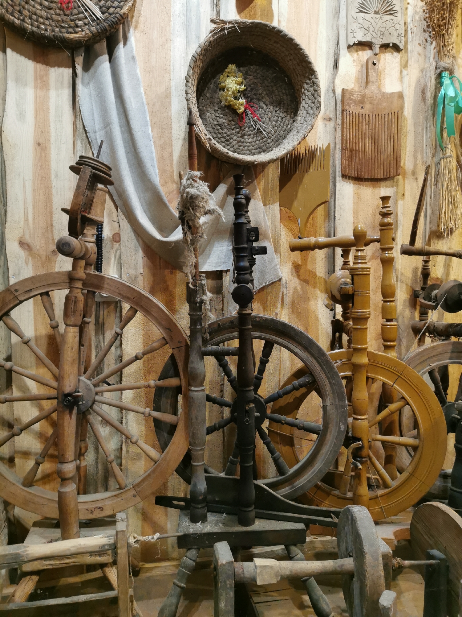 The image displays a collection of antique spinning wheels and weaving tools displayed against a rustic wooden wall. The items appear to be arranged as an exhibit, possibly in a museum or a workshop, showcasing traditional textile production methods. In the upper left and right corners, two woven baskets are visible, with one containing dried flowers. Several wooden combs, likely for wool or flax processing, are also hung on the wall, along with a bundle of dried plants tied with a green ribbon. The spinning wheels vary in size and design, some made of dark wood and others of lighter, possibly varnished wood. A piece of rough, unspun fiber is attached to one of the spinning wheels, suggesting it's in a state of use or demonstration. The overall impression is one of historical preservation and traditional craftsmanship. This scene is located in Шчарчоўскі сельскі Савет, Belarus.