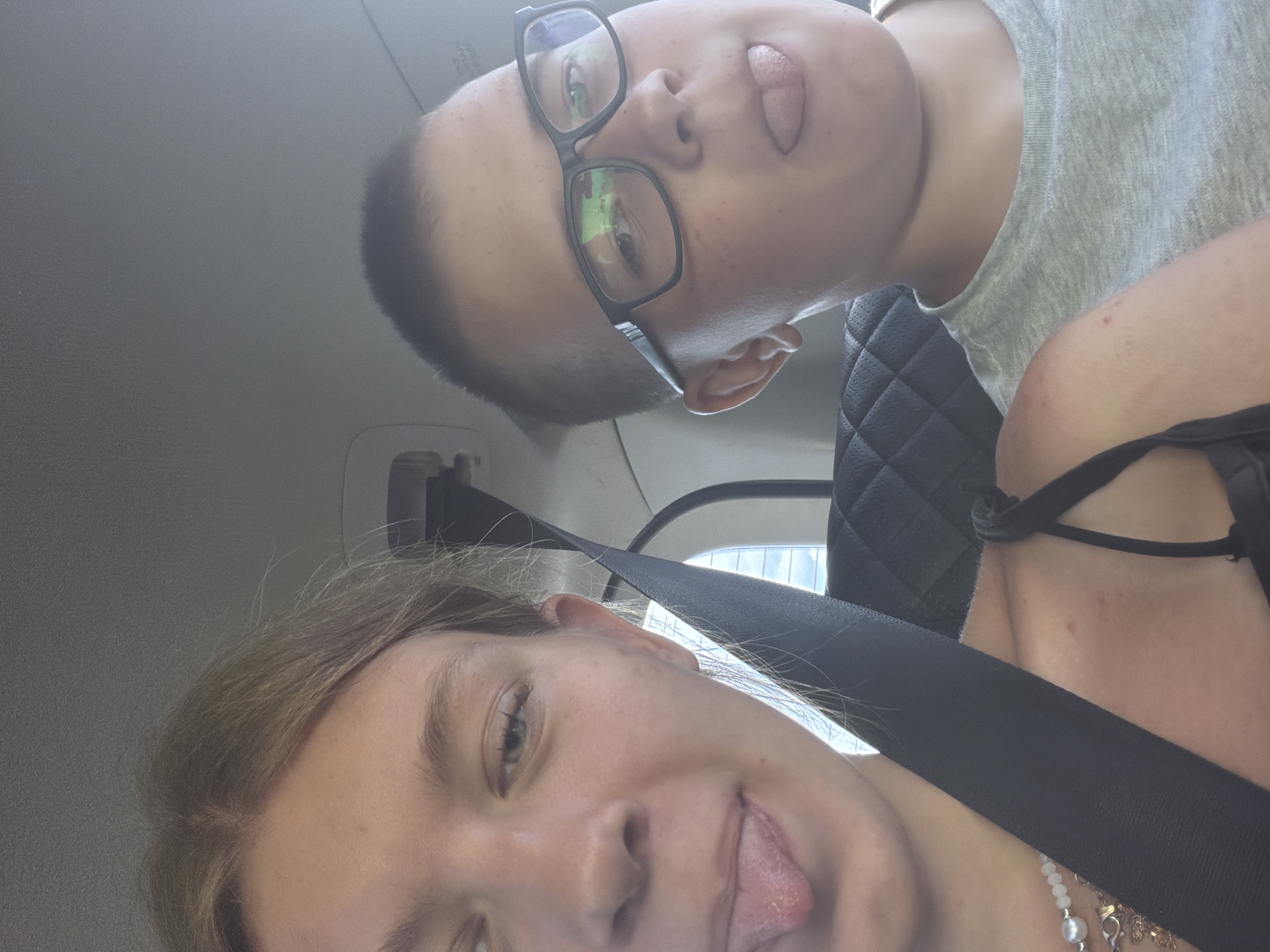 Two children, a girl and a boy, are seated in a car. The girl, on the left, has blonde hair and is sticking her tongue out. She is wearing a necklace with pearls and a gold pendant. The boy, on the right, wears glasses and is also sticking his tongue out. He is wearing a grey t-shirt. They appear to be in the back seat of a car, as suggested by the seatbelt and car interior visible. The background shows a blurred view of what might be a building and sky, possibly indicating the car is moving or parked near a window. The location is identified as Novoe Feizullovo, Russia.