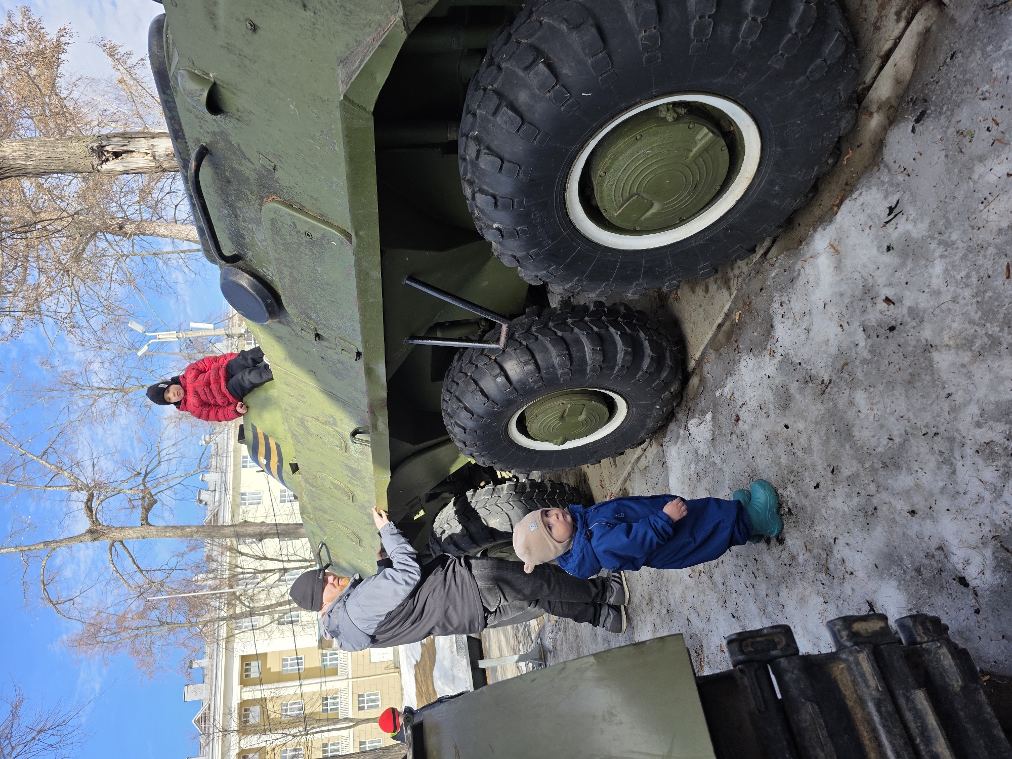 The scene is set outdoors in Votkinsk, Russia, during what appears to be late winter or early spring, given the patchy snow and bare deciduous trees. A clear blue sky is visible overhead.
In the center of the image, a large, olive-green wheeled armored personnel carrier (APC) or infantry fighting vehicle (IFV) with prominent black tires and white rims is displayed. On the ground next to this vehicle stands an adult male, wearing a grey and black hooded jacket, dark pants, and a black beanie, with a reddish beard. He is positioned with his hands on the upper side of the vehicle's hull, appearing to interact with it.
Seated on the top-front section of the same military vehicle is an older child, dressed in a red puffer jacket and a black beanie, looking towards the left. In the immediate foreground, a toddler in a blue full-body snowsuit, a light brown beanie with earflaps, and teal boots stands on the snowy and icy ground, facing towards the right.
To the far left of the frame, a portion of a second military vehicle, featuring a dark tracked system, is partially visible. The background features a multi-story building with a light yellow facade, white columns, and multiple windows, partially obscured by bare trees.