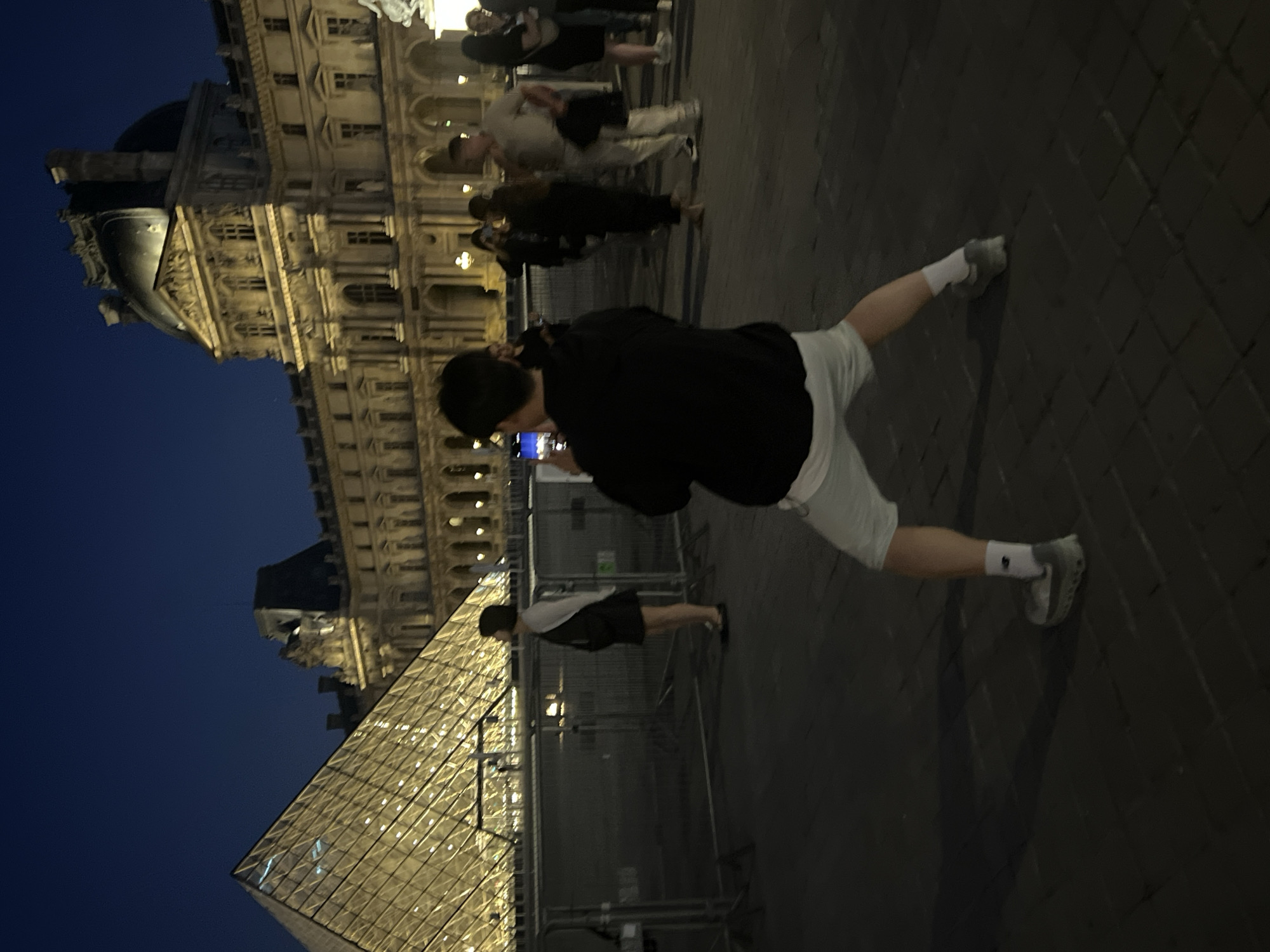 The image depicts the exterior of the Louvre Museum in Paris, France, at dusk. A large glass pyramid, illuminated from within, is visible on the left side of the frame. Behind the pyramid, the ornate facade of the museum building is lit up, showcasing its classical architecture.
In the foreground, a young person wearing a black hooded sweatshirt, light-colored shorts, white socks, and grey sneakers is facing away from the camera, holding a smartphone and appearing to take a photograph. Their stance is wide and dynamic, with their legs bent and spread apart.
Several other individuals are visible in the background, some standing near the pyramid and others further back in front of the museum. They are mostly out of focus. Security barriers are present between the pyramid and the people in the background. The ground is paved with cobblestones. The sky above is a deep, dark blue, indicating nighttime.