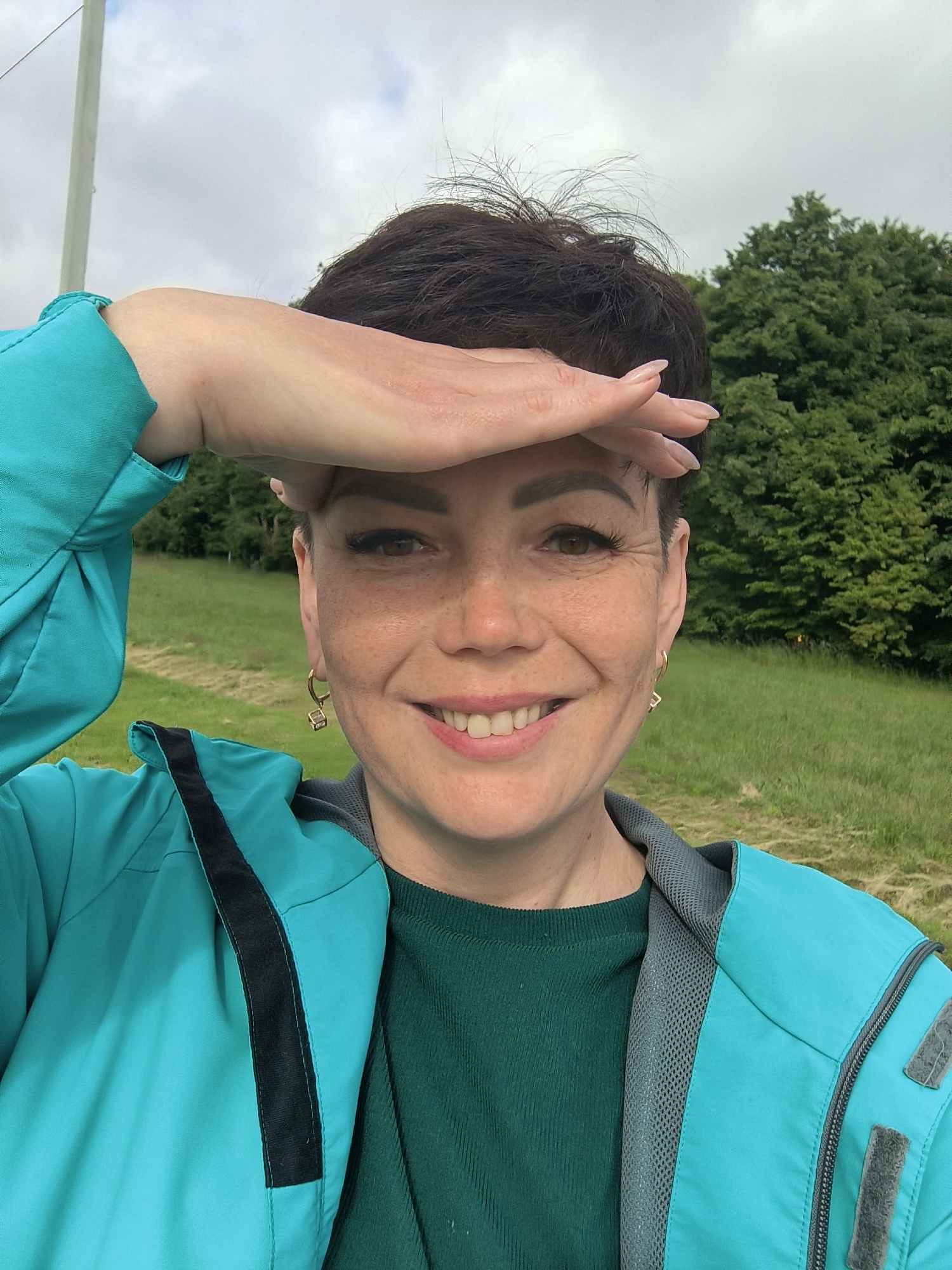 A woman with short, dark hair and freckles is taking a selfie outdoors. She is wearing a teal jacket over a dark green top and has her right hand raised to shield her eyes from the sun. Her expression is a smile. The background shows a grassy field with trees and a cloudy sky. There is a white pole on the left side of the image. The image is not of the city of Laskino, Russia.