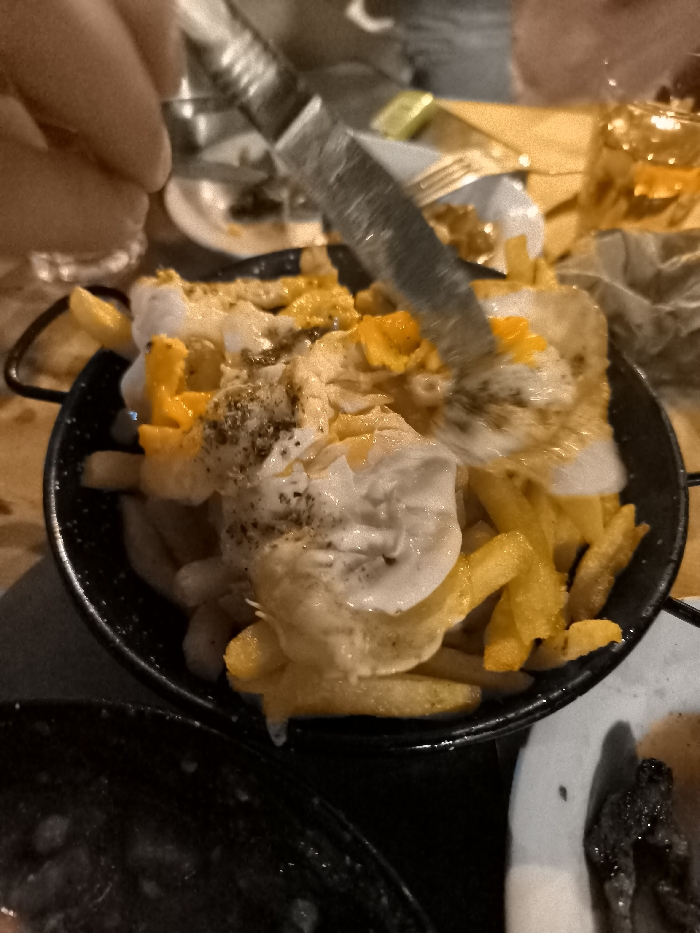 The image captures a close-up view of a person cutting into a dish of food at what appears to be a restaurant or bar in Nea Erithrea, Greece. The main subject is a black, round skillet or bowl filled with golden-brown french fries, generously topped with multiple fried eggs, one with a visible bright orange yolk, along with a creamy white sauce or dressing and dark seasoning, likely pepper or herbs.
A person's hand is visible on the left side, holding a metallic knife, actively slicing into one of the fried eggs. In the background, elements of a dining setting are blurred, including a glass with a clear liquid, a small white plate with food remnants, and a fork. To the right, part of a glass containing an amber-colored drink and some crumpled paper are visible. Below the main dish, another dark-colored dish containing a stew-like food is partially seen.
The scene depicts a social dining event, likely during the evening or night, indicated by the dim, warm, artificial lighting. The activity is the act of serving or eating this hearty dish. No text is visible within the image. The focus is on the food and the immediate action of preparation or consumption.