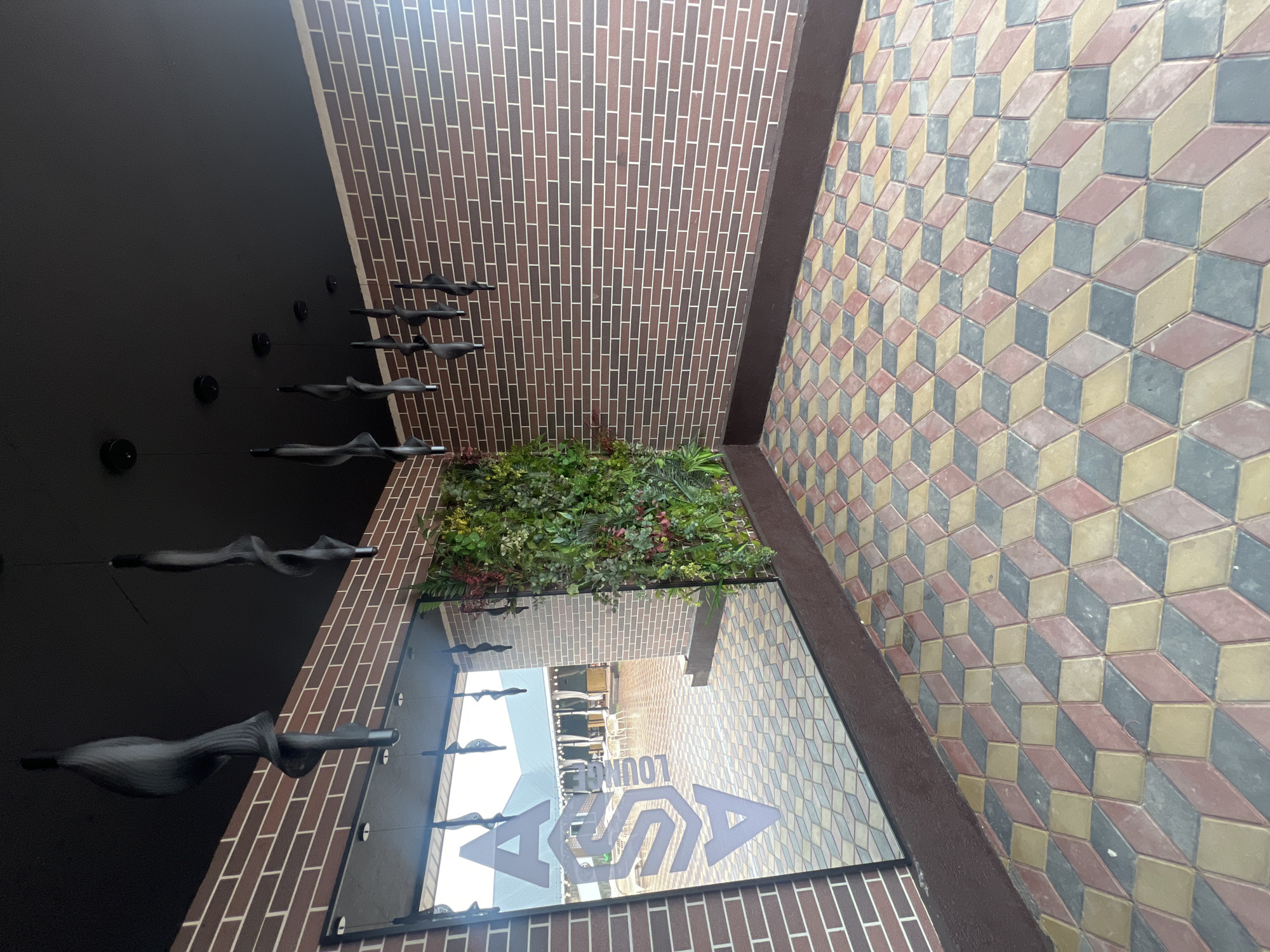 The image shows an outdoor seating area with a brick wall, a vertical garden, and a mirrored wall with a logo. The floor is paved with colorful interlocking bricks arranged in a geometric pattern that creates an optical illusion of depth. There are several black, twisted hanging light fixtures from the ceiling. The mirrored wall reflects an outdoor seating area with tables and chairs, suggesting the location is a cafe or restaurant. The text on the mirrored wall appears to read "ASA LOUNGE." The overall aesthetic is modern and stylish, with a focus on natural elements like the vertical garden. The location is specified as Ақши, Kazakhstan.