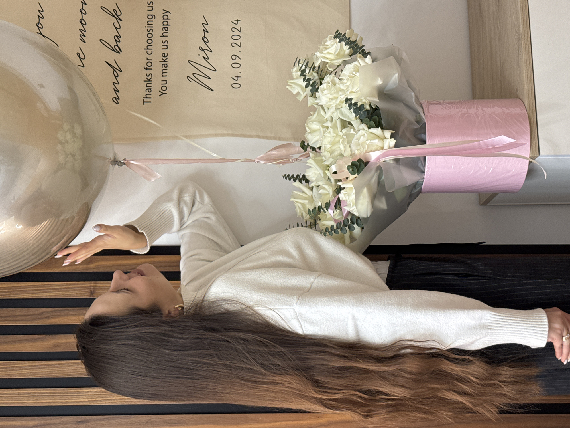 A photograph shows a young woman with long brown hair lying on what appears to be a wooden floor with black accents. She is wearing a white sweater and dark-colored pants. Next to her are a bouquet of white roses in a pink container and a large transparent balloon. Behind the balloon and flowers is a beige sign with the text "Thanks for choosing us. You make us happy Miron 04.09.2024." The setting appears to be indoors. There is no explicit information or detail provided in the image that reveals the specific location in Akulovo (Gp Kubinka), Russia.