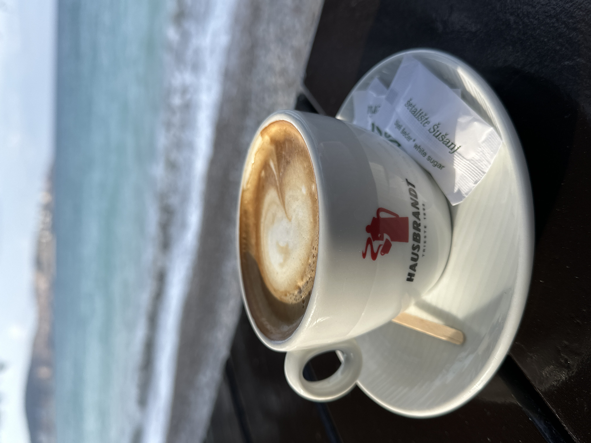 A close-up, high-angle shot depicts a white ceramic coffee cup containing a beverage with visible latte art, centrally positioned on a matching saucer. The cup features a red logo of a steaming moka pot above "HAUSBRANDT TRIESTE 1892". A small, light-colored wooden stirrer rests on the saucer, alongside two white sugar packets. One packet is clearly visible and reads "šetaliste Šušanj" and "bijeli šećer | white sugar". The cup and saucer are situated on a dark, textured surface, likely a table. The background, out of focus, reveals a body of water with light blue-green hues and white wave crests approaching a shore. Beyond the water, indistinct landmasses or hills are visible under a pale sky. The presence of the "šetaliste Šušanj" sugar packet indicates the scene is located on a promenade or boardwalk in the city of Šušanj, Montenegro.