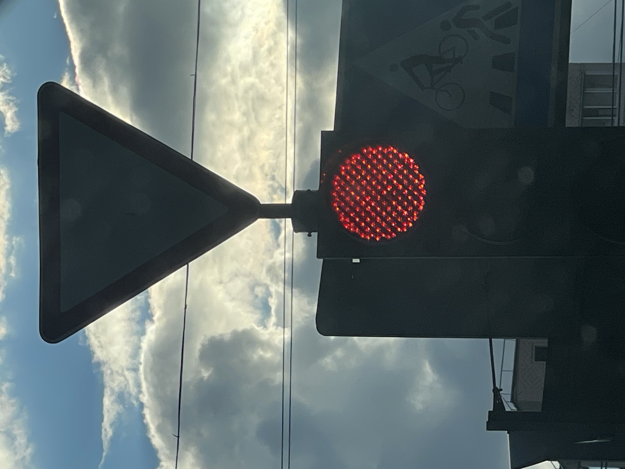 The image captures a traffic light with a red illuminated circular display, positioned below an inverted triangular "Give Way" sign. To the right of the traffic light, a blue rectangular sign displays silhouettes of a cyclist and a pedestrian on a crosswalk. Overhead, power lines traverse the frame against a cloudy sky. In the background, elements of urban architecture, including a building with windows, are visible. The scene appears to be a street in Minsk, Belarus, possibly near a crosswalk and intersection.