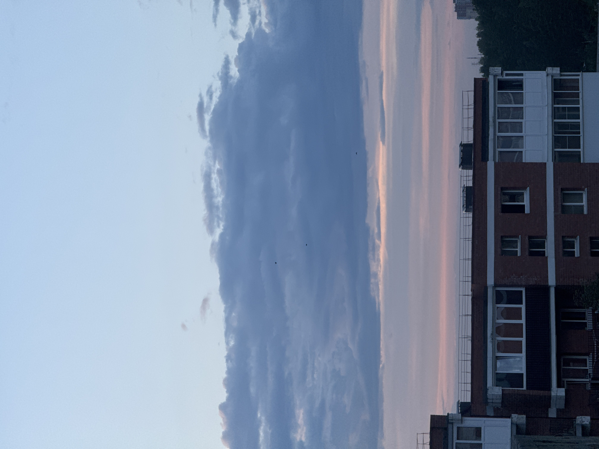 The image depicts a twilight sky over a multi-story residential building in Markova, Russia. The sky is a gradient of pale blue at the top, transitioning to shades of grey and purple with scattered clouds. Below the sky, the upper floors of the brick apartment building are visible. The building features a red brick facade with numerous windows, some of which are dark and appear empty, while others have white window coverings or glass balconies. A railing runs along the rooftop of the building. To the right of the building, a dense line of dark green trees is visible, and in the far distance, another tall residential building can be seen. The overall scene suggests a quiet urban evening.