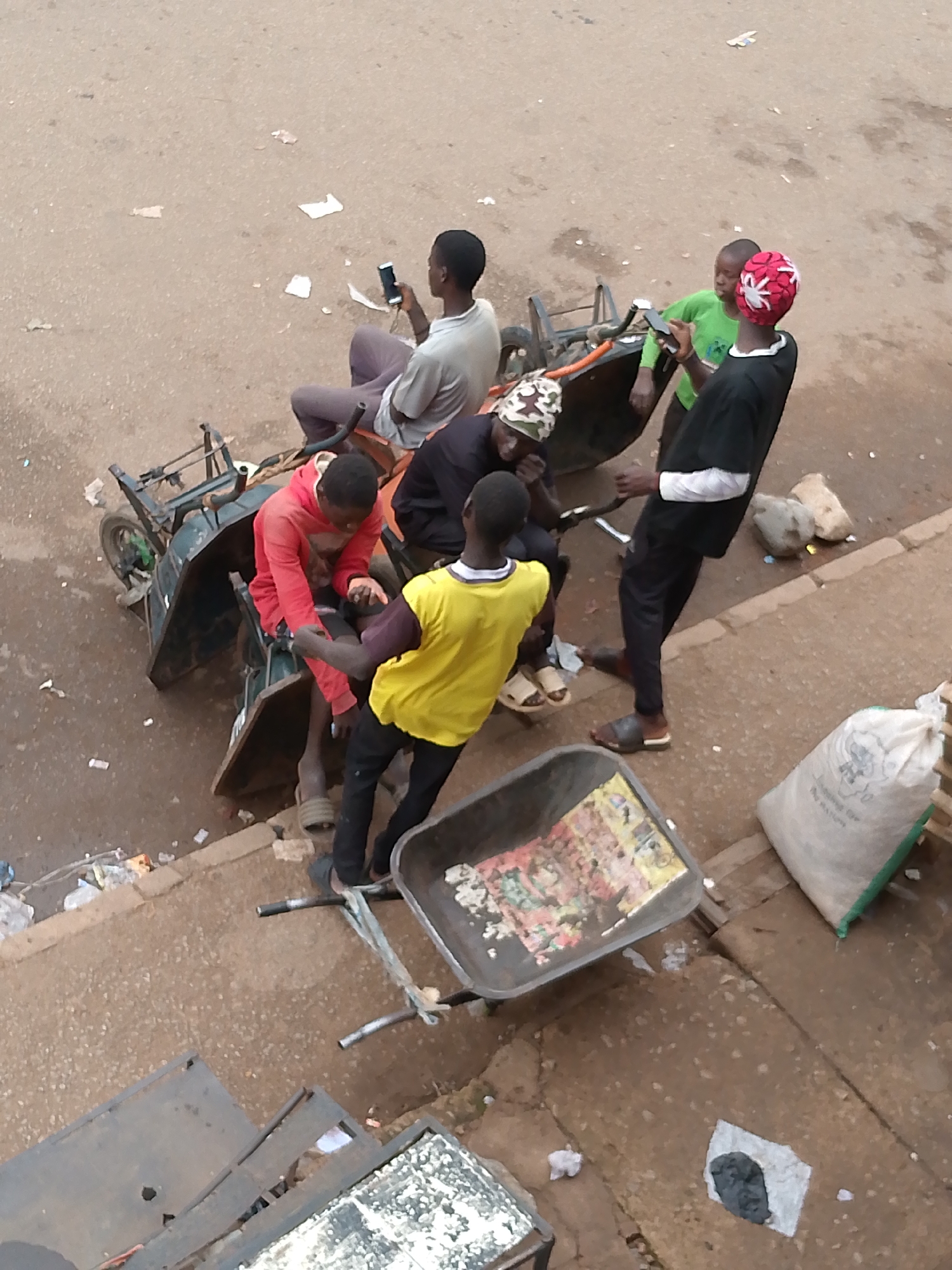 The image shows a street scene in Bukuru, Nigeria. Six individuals are gathered around an overturned motorized cart or tricycle, and a wheelbarrow is nearby.
One person, wearing a red shirt, is seated inside the cart, supported by another individual in a yellow shirt. A third person is sitting on the frame of the vehicle. One person is standing and looking at their mobile phone, wearing a green shirt and a red-and-white patterned head covering. Another person is standing nearby wearing black clothing. The final person is sitting on the cart and looking at their mobile phone, wearing a gray shirt.
A white bag with green trimming sits on the ground beside the group. There are assorted litter and scraps of paper scattered on the road and the sidewalk, including some items on top of the wheelbarrow. The background is a paved road with buildings and other urban elements visible.