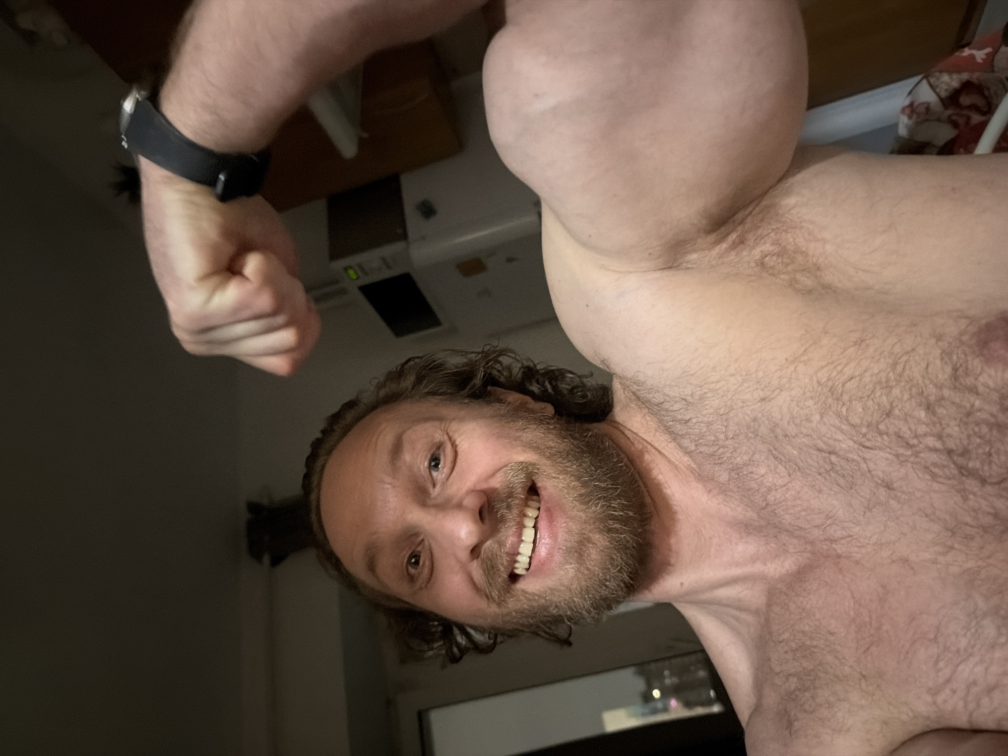 A shirtless man with a beard and long hair is posing and smiling at the camera. He is flexing his arm, which has a black watch on the wrist. The background is dark, but a white appliance and a window with blurred lights can be seen. The location is impossible to determine from this image.