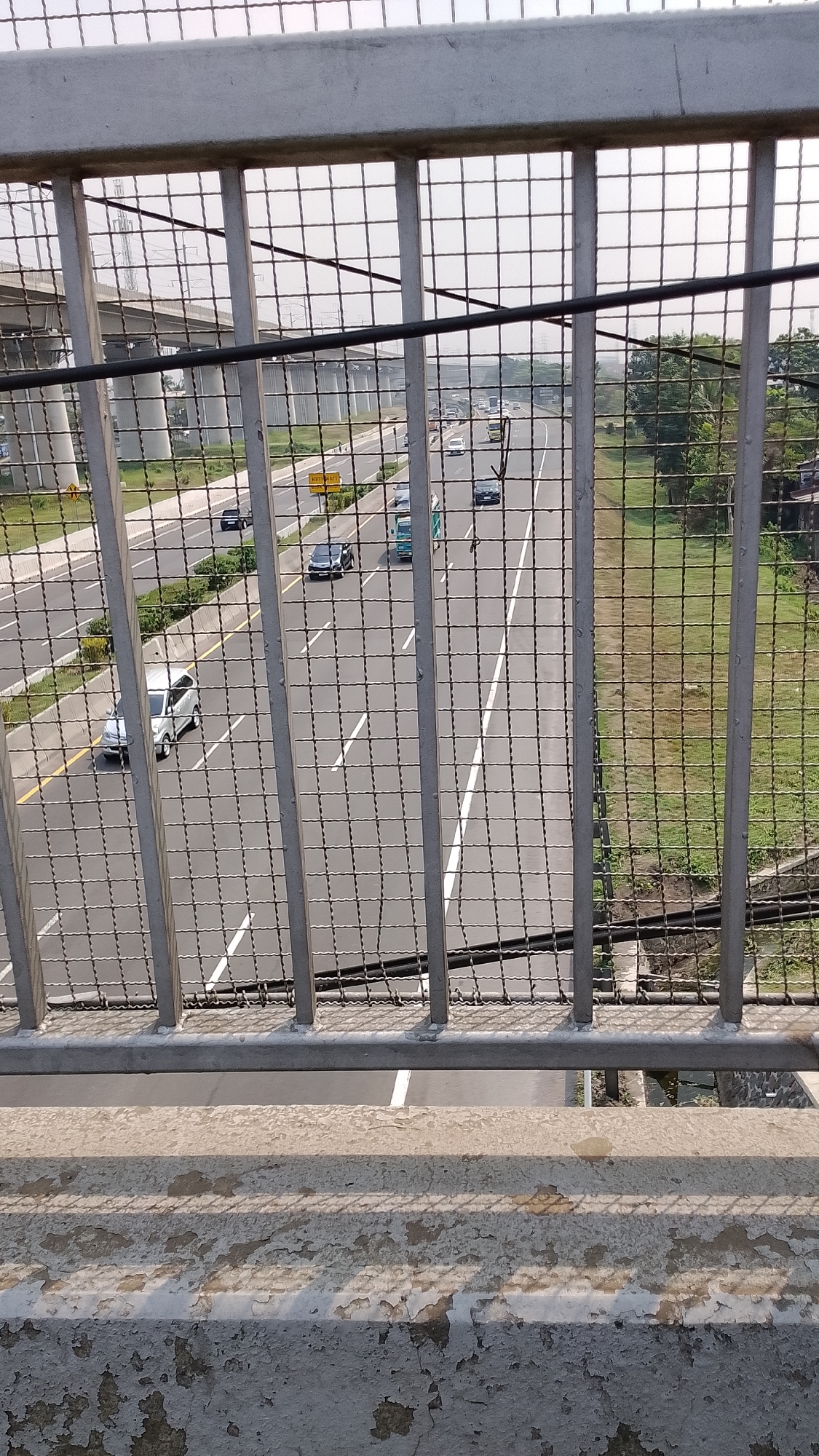 The image shows an elevated view of a multi-lane highway, likely an expressway, seen through a metal fence. The highway has multiple lanes of traffic with vehicles including cars and a bus. The highway is flanked by green grassy areas and some trees on the right. On the left, an elevated structure, possibly another highway or a bridge, is visible. The foreground consists of a concrete barrier with peeling paint, and the metal fence that obstructs the view. The sky is overcast. This scene is consistent with an urban highway environment found in Bandung City, Indonesia, a region with extensive highway infrastructure.
