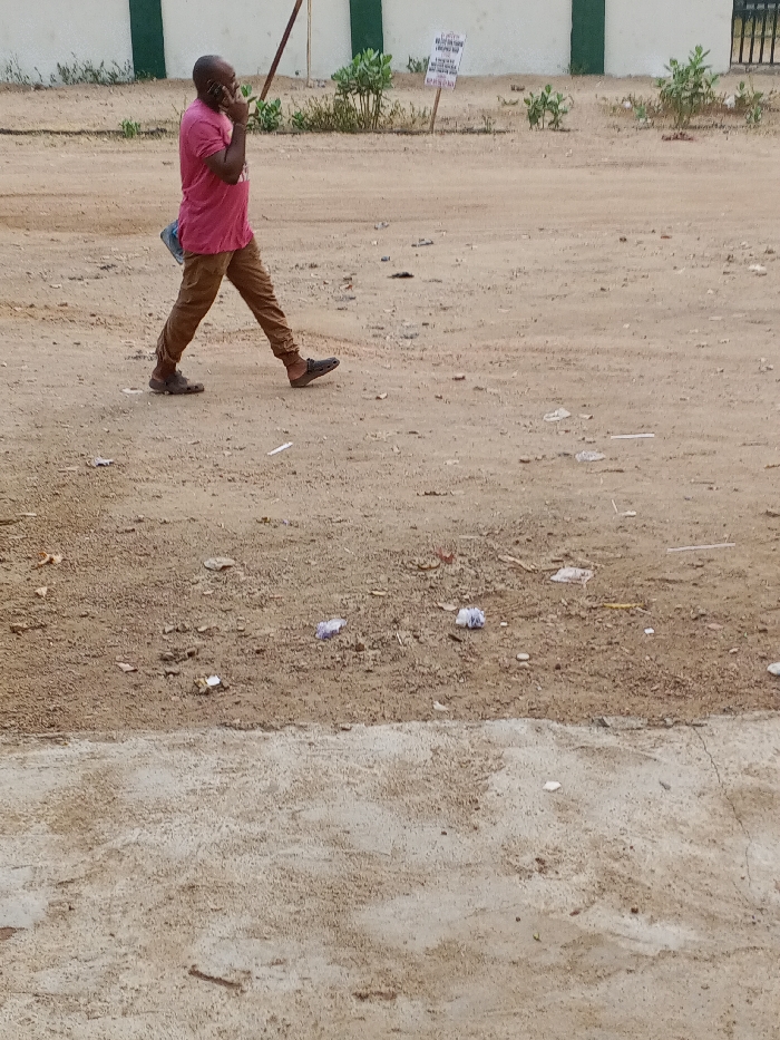 A man in a pink shirt and brown pants walks across a dirt road while talking on a cell phone. The ground is littered with trash and debris. In the background, there's a white wall with green accents and some vegetation. A sign with text on it is visible near the wall. The foreground features a lower concrete surface.