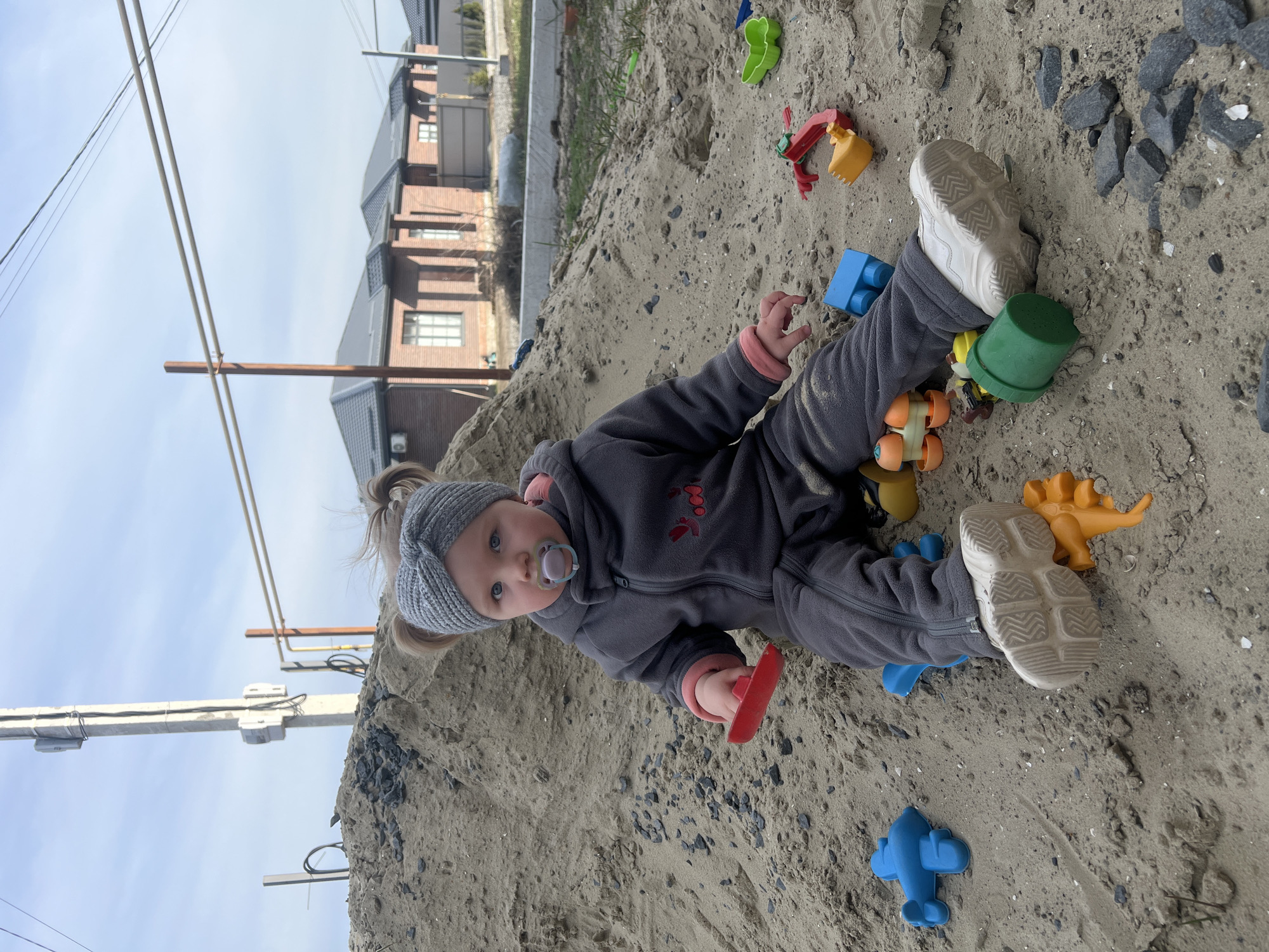 A young child, estimated to be a toddler, is seated directly on a large pile of light brown sand, facing forward towards the viewer. The child wears a grey knitted turban-style headband with hair gathered into two small buns, a grey hooded fleece jumpsuit with a visible pink lining at the cuffs, and white athletic shoes with textured soles. A blue and purple pacifier is present in the child's mouth. The child holds a small red plastic shovel in its right hand. Scattered around the child on the sand are multiple colorful plastic toys, including a blue airplane, a green bucket, an orange wheeled toy, an orange animal figure, and a red and yellow excavator-like toy. Dark grey, angular stones are dispersed within the sand, particularly to the child's right. In the background, under a clear light blue sky, several brick houses with dark grey tiled roofs and white-framed windows are visible, indicative of a modern residential development. A tall concrete utility pole equipped with electrical meters and wires stands prominently to the left of the background structures, alongside a wooden pole supporting horizontal utility pipes to the right. The scene takes place outdoors in what appears to be a developing residential area in Leninakan, Russia.