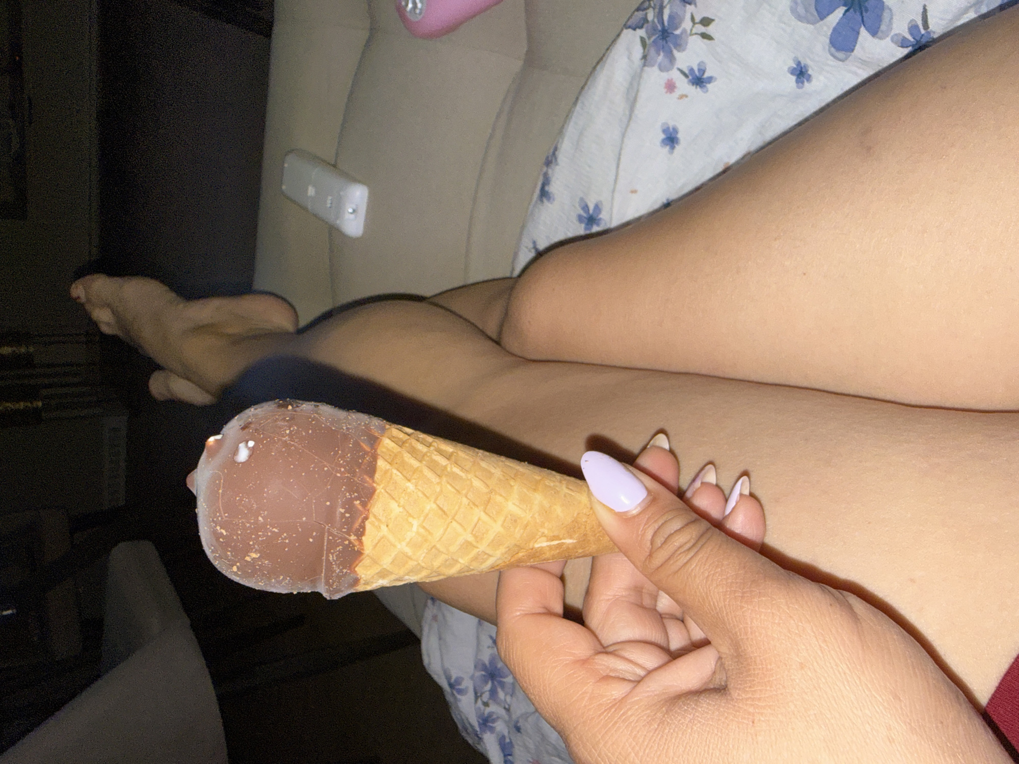The image captures a close-up, low-light indoor scene featuring a person holding an ice cream cone while relaxing. The person, whose bare legs and feet are visible, is seated on what appears to be a light-colored couch or bed, partially covered by a white fabric with blue floral patterns.
In the foreground, a hand with light purple manicured nails holds an ice cream cone. The cone itself is a classic waffle cone, topped with a chocolate-dipped ice cream that has small white particles, possibly sprinkles or nuts, adhering to its surface.
In the background, several objects are discernible. A white remote control, resembling a Nintendo Wii remote, is positioned on the couch near the person's legs. Further back and slightly out of focus, a pink electronic device with a rounded shape is visible. The overall lighting suggests it is evening or nighttime. No discernible text is visible on any of the objects. The setting appears to be a cozy, personal space like a living room or bedroom.