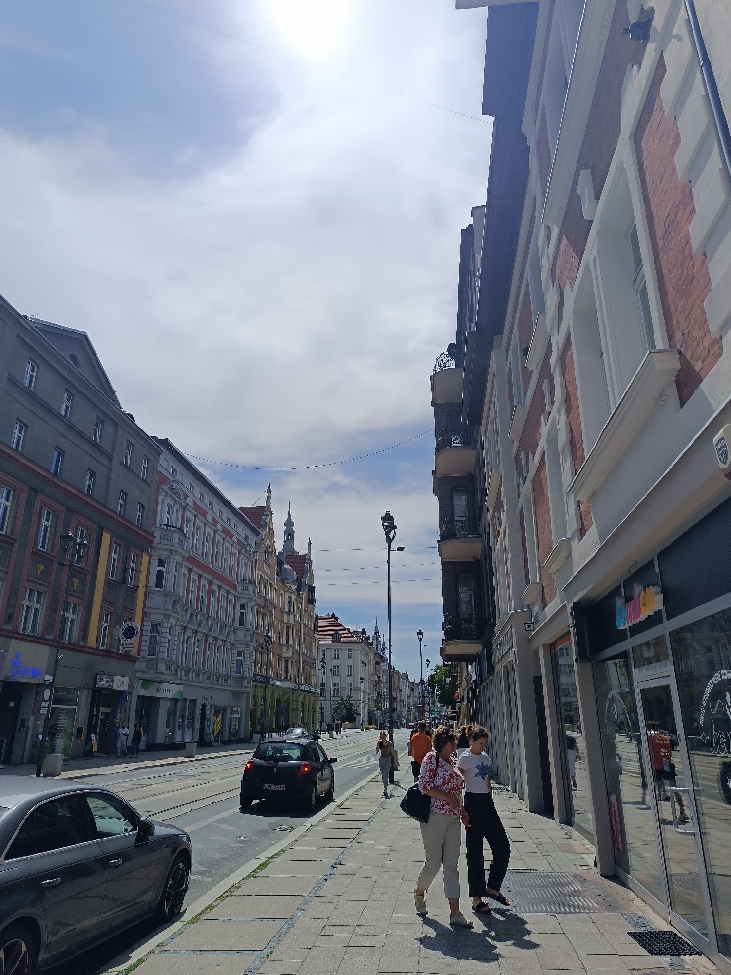 In Gliwice, Poland, this image captures a street scene on a sunny day. A row of buildings lines the right side of the street, featuring red brick and white architectural details. On the left, a different architectural style is visible, with grey facades and ornate window frames. People are walking on the sidewalk, some appearing to window shop, while others are in transit. Several cars are present, including a dark-colored sedan in the foreground and a black hatchback further down the street, which is driving on the road. The sky is partly cloudy. The environment suggests a bustling urban area with a blend of historical and modern elements.