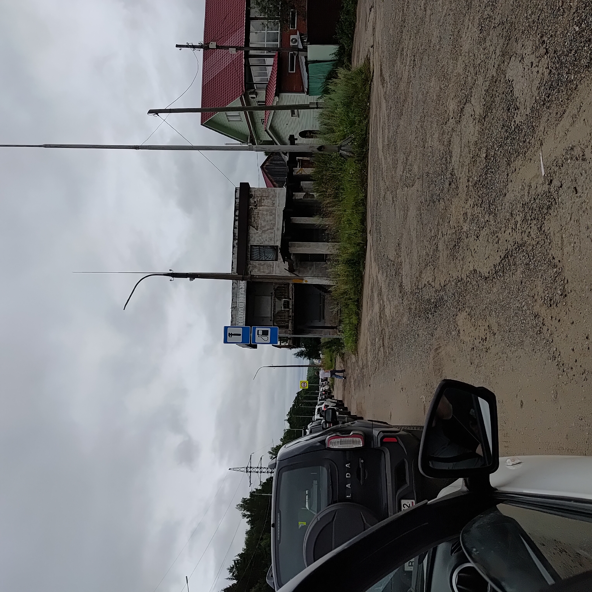 The image captures a traffic jam on a dirt road in Chaikovsky, Russia, under an overcast sky. A line of cars stretches into the distance, indicating a standstill. In the foreground, the rear of a black Lada SUV is visible, with its spare tire mounted on the back. To the right, an abandoned, dilapidated concrete building stands with overgrown vegetation around its base. A sign depicting a hammer and a gas pump is affixed to a pole in front of this building. Further down the road, a man in a white t-shirt and blue jeans walks away from the camera. In the background, several houses with red and green roofs are visible, along with power lines and trees. The road surface is rough and uneven, composed of dirt and gravel.