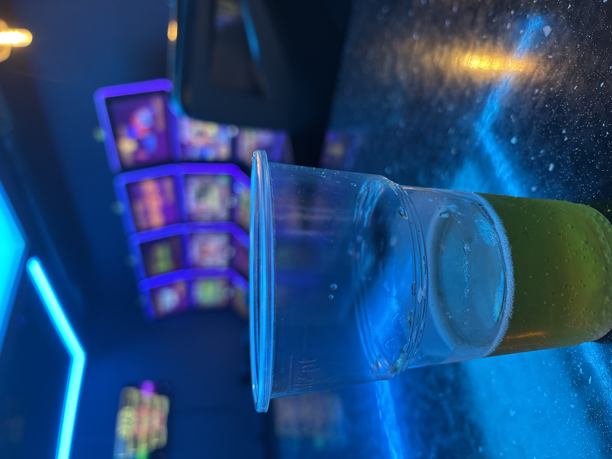 A clear plastic cup, partially filled with a yellow liquid, rests on a dark, reflective surface illuminated with blue and yellow light. Condensation is visible on the cup's exterior. In the background, out-of-focus, there are illuminated screens suggesting gaming machines or displays. A blue neon light is visible to the left. The precise location within the city of Pleven, Bulgaria, cannot be determined from the image.