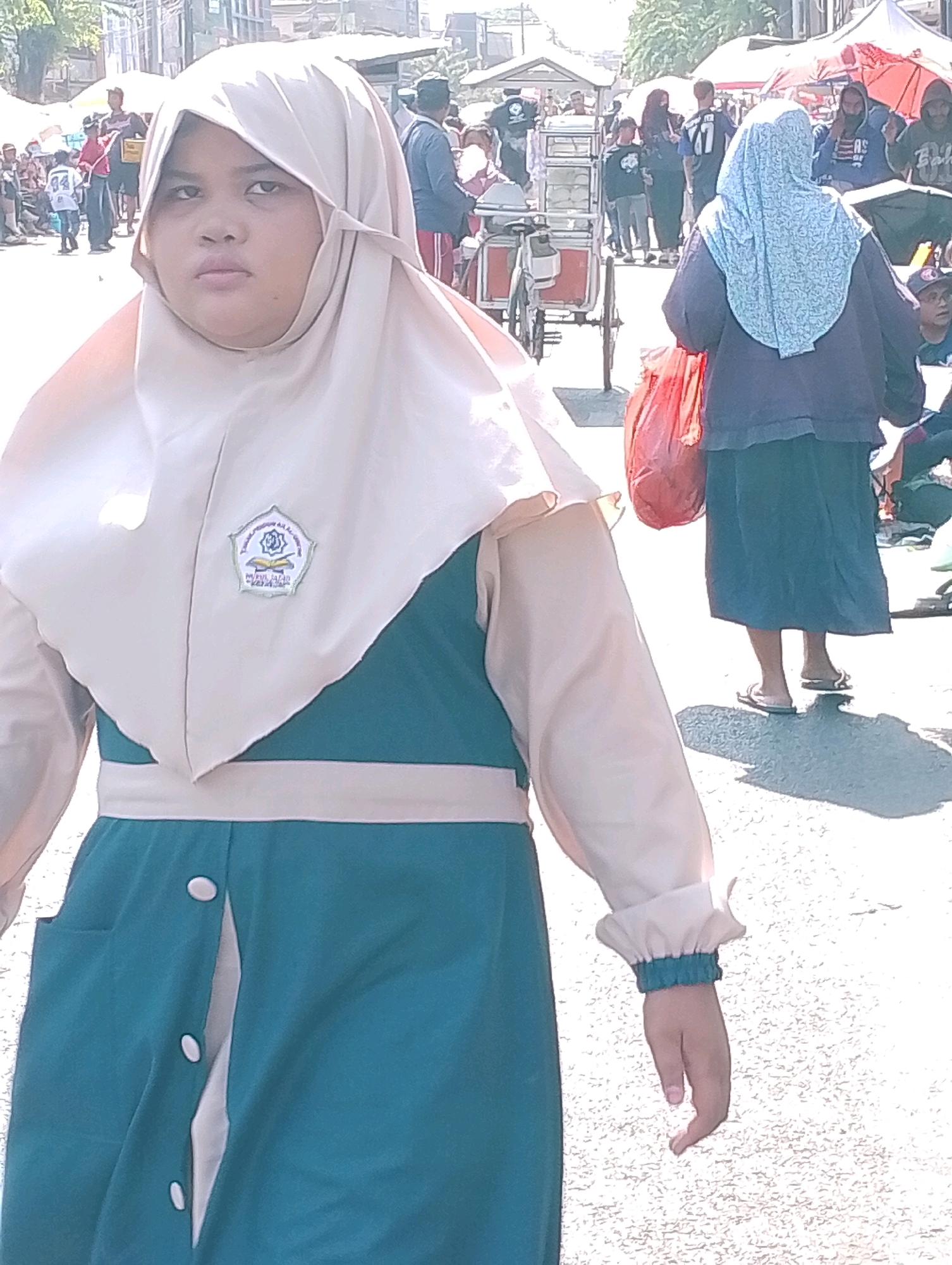 A young person wearing a light beige hijab and a teal and beige dress stands in the foreground, looking off to the left. The dress features a beige band at the waist and a beige collar. A pentagonal emblem is visible on the left side of the hijab. In the background, a bustling street scene unfolds with tents, market stalls, and a crowd of people. A food cart with stacked items is visible on the left, and a person in a blue hijab and dark clothing walks by carrying a red bag. The setting appears to be an outdoor market or festival, likely in the city of Probolinggo, Indonesia, given the cultural attire and general ambiance. The ground is paved with asphalt. The lighting suggests it is daytime, with sunlight casting shadows.
