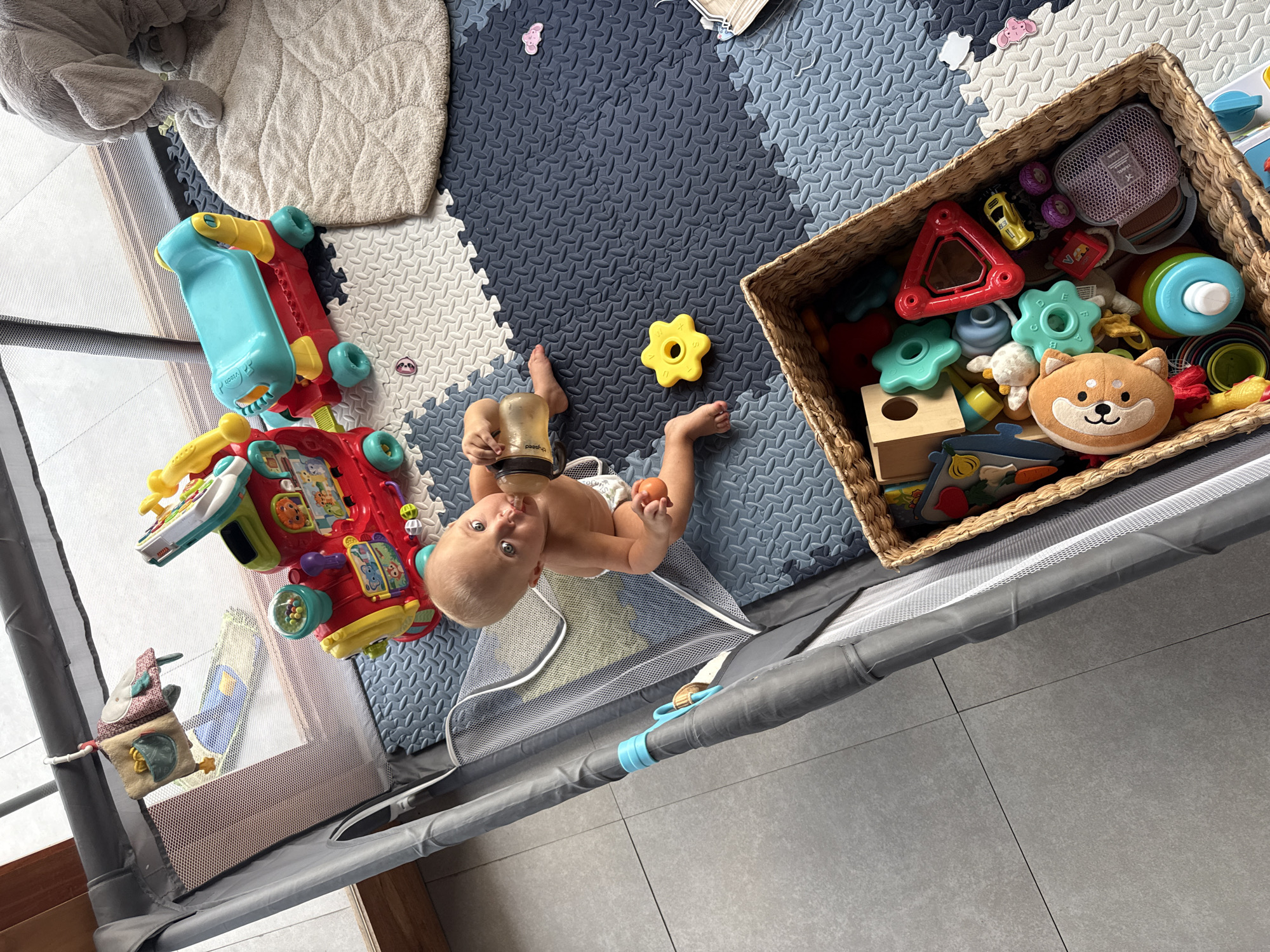 An overhead shot captures a baby lying on their back in an indoor play area in Mengwi, Indonesia. The baby, wearing only a diaper, holds a "pigeon" brand bottle to their mouth with one hand, while clutching a small orange toy in the other. Their blue eyes gaze directly at the camera.
The baby is situated on a multi-colored foam playmat with interlocking tiles in dark blue, light blue, and white patterns. Surrounding the playmat is a grey fabric and mesh playpen enclosure, beyond which a grey tiled floor is visible.
Numerous toys are scattered around the play space. To the baby's left is a red and blue "VTech" baby walker or push toy. A yellow star-shaped toy rests on the dark blue playmat near the baby's feet. To the baby's right, a woven wicker basket overflows with a variety of toys, including a brown shiba inu plush, wooden blocks, colorful plastic shapes (some marked with letters like "E" and "B"), stacking rings, small vehicles, and a "WABA" branded mesh bag. A soft grey blanket or mat is partially visible in the upper left corner of the playmat. A small fabric soft book hangs from the playpen structure on the left side.
The scene depicts a calm, daytime moment of a baby playing and feeding within a safe, designated play area.