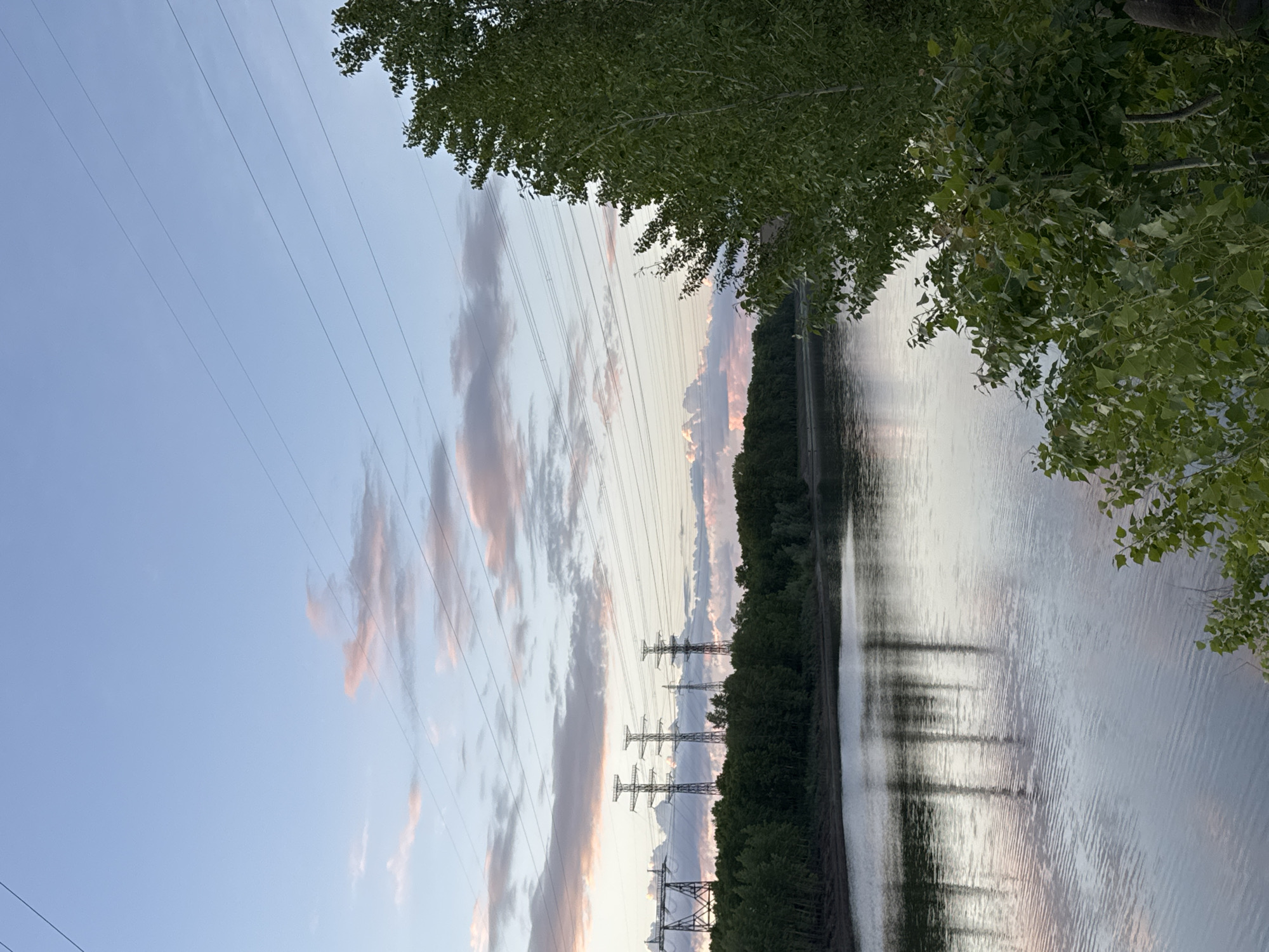 The image captures a serene landscape at what appears to be dusk or dawn, showcasing a wide river, dense foliage, and electrical power infrastructure under a softly lit sky.
The sky, occupying the upper left portion of the frame, is a light blue, adorned with scattered, elongated clouds that exhibit subtle pink and orange hues, indicative of the setting or rising sun. Several thin, dark power lines crisscross the sky from the upper left to the lower right.
In the upper right, the dark green leaves of a tree hang into the frame, partially obscuring the view. Below this foliage and extending across the lower half of the image is a broad body of water, likely a river. Its surface is relatively calm, displaying gentle ripples and reflecting the muted sky and the silhouettes of distant features.
Along the far bank of the river, a continuous line of dense, dark green trees creates a natural barrier. Interspersed within or behind these trees are five prominent electrical transmission towers (pylons) with complex metallic structures, stretching horizontally across the middle ground. These towers stand as dark, angular silhouettes against the brighter sky behind them.
The scene combines natural elements with industrial infrastructure, bathed in the tranquil, soft light of the transitional hours of the day. There is no visible human activity or text. The location, Chaikovsky, Russia, suggests a view typical of a river landscape in that region.