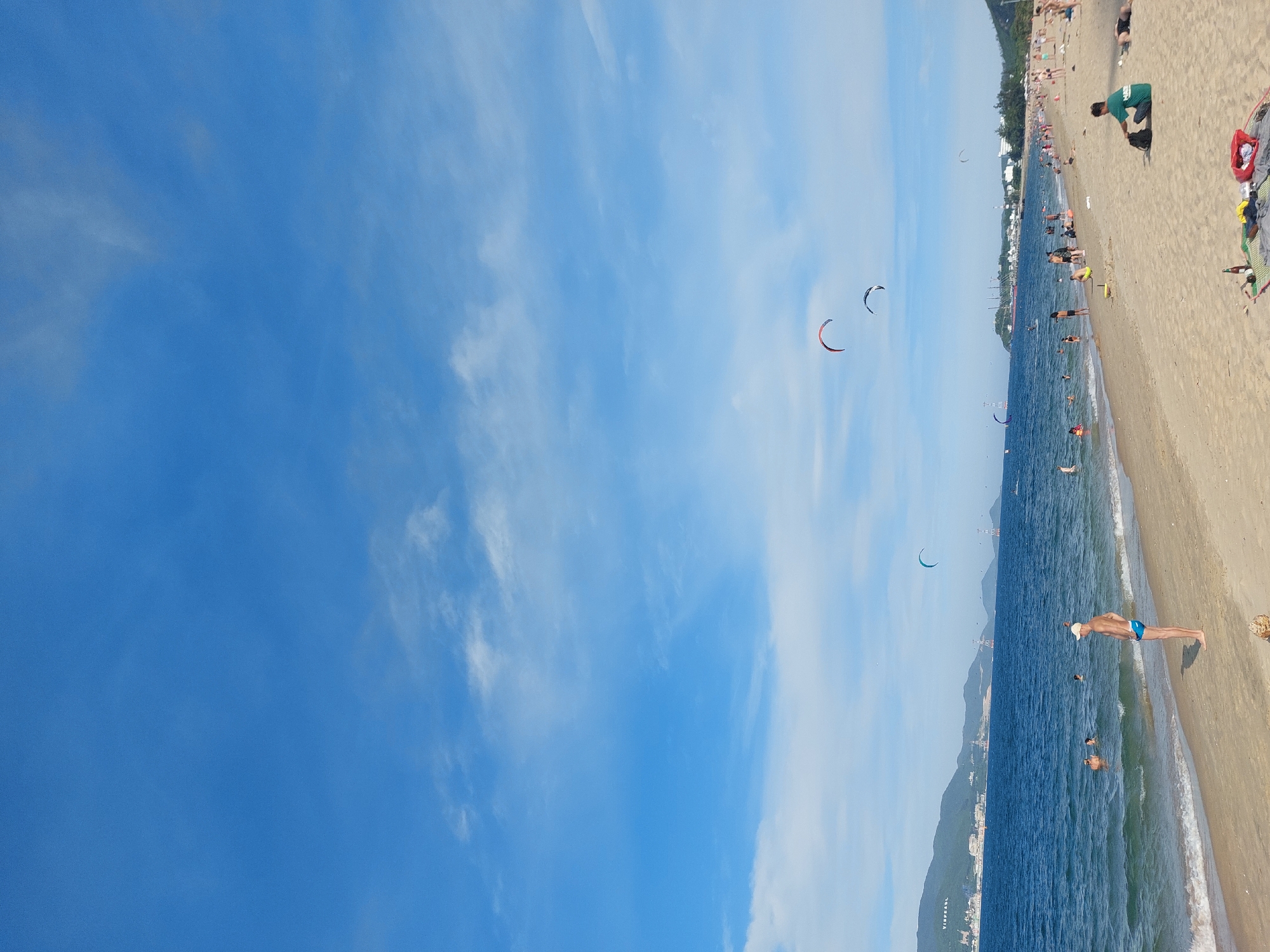 This is a wide-angle, eye-level shot of a beach scene in Nha Trang Ward, Vietnam. The sky is a vibrant blue with scattered white clouds. The ocean is a deep blue, with gentle waves reaching the sandy shore. Several kite surfers are visible in the water, their colorful kites creating arcs against the sky.
On the beach, there are numerous people enjoying the day. Some are swimming or wading in the water, while others are sunbathing, sitting, or walking along the sand. A man in blue swim trunks and a white cap stands near the water's edge, facing away from the camera. Further down the beach, a person in a green shirt sits on the sand with a backpack.
The background features a coastline with hills and mountains covered in greenery. A city or town is visible along the coast, with buildings and infrastructure such as cranes and power lines. The overall impression is a busy and lively beach on a sunny day.