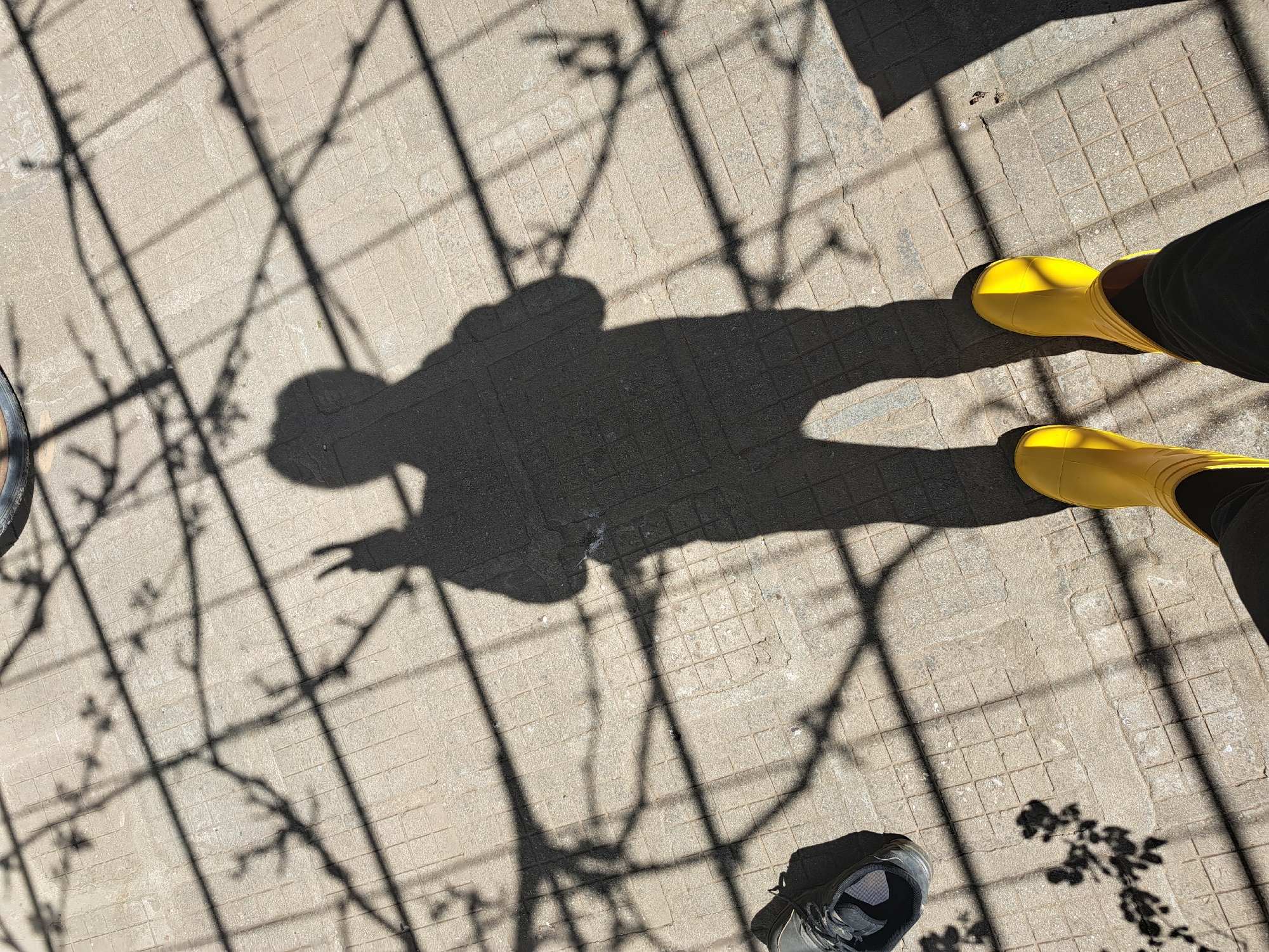 The image shows a shadow of a person with their left hand making a peace sign. The person is wearing yellow boots. Another shoe, gray and black, is visible in the bottom of the frame. A pattern of shadows from a fence and foliage are visible on the tiled ground. The location is Unknown, Unknown.