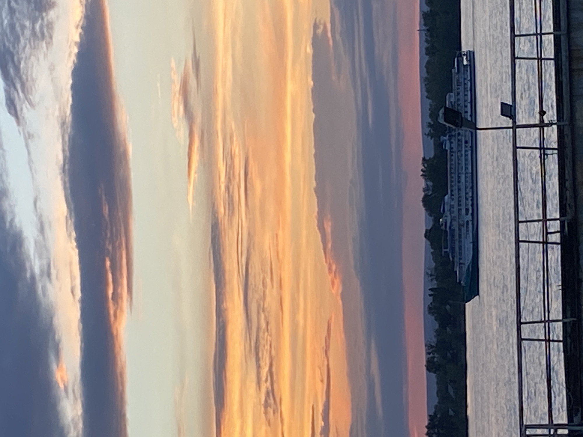 This is a vertical, medium-shot photograph capturing a sunset over a body of water, likely a river, with a docked boat and a pier in the foreground. The sky is filled with dramatic clouds painted in hues of orange, yellow, and blue, indicative of a sunset. The sun's rays are diffused, creating a soft, ethereal glow across the sky.
A large, multi-deck boat is moored alongside a pier on the right side of the frame. The boat is predominantly white with several decks and appears to be a passenger vessel. The pier is a dark, metallic structure with a grid-like pattern, suggesting it's a functional part of the waterfront. The water itself is calm, reflecting the colors of the sky, with ripples visible on its surface.
On the far left of the image, a silhouetted shoreline with trees can be seen against the twilight sky. The time of day is clearly sunset, given the colors and lighting. The weather appears to be clear, allowing for a vibrant sunset display. The location context of Novokuybyshevsk, Russia, places this scene in a specific geographical setting, though no specific landmarks within the city are identifiable from the image itself. There is no visible text in the photograph. The overall mood is serene and picturesque, evoking a sense of calm at the end of the day.